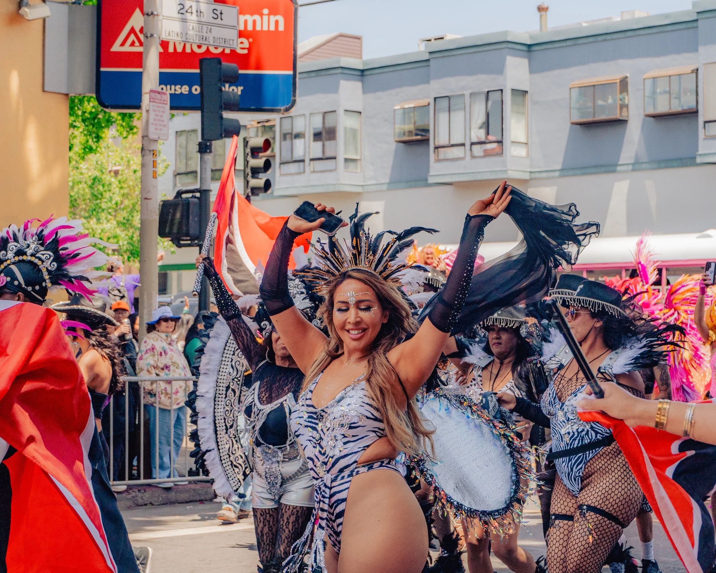 Woman dancing in a parade wearing a zebra print costume with feathers and face paint, surrounded by other parade participants in colorful costumes and feathers.