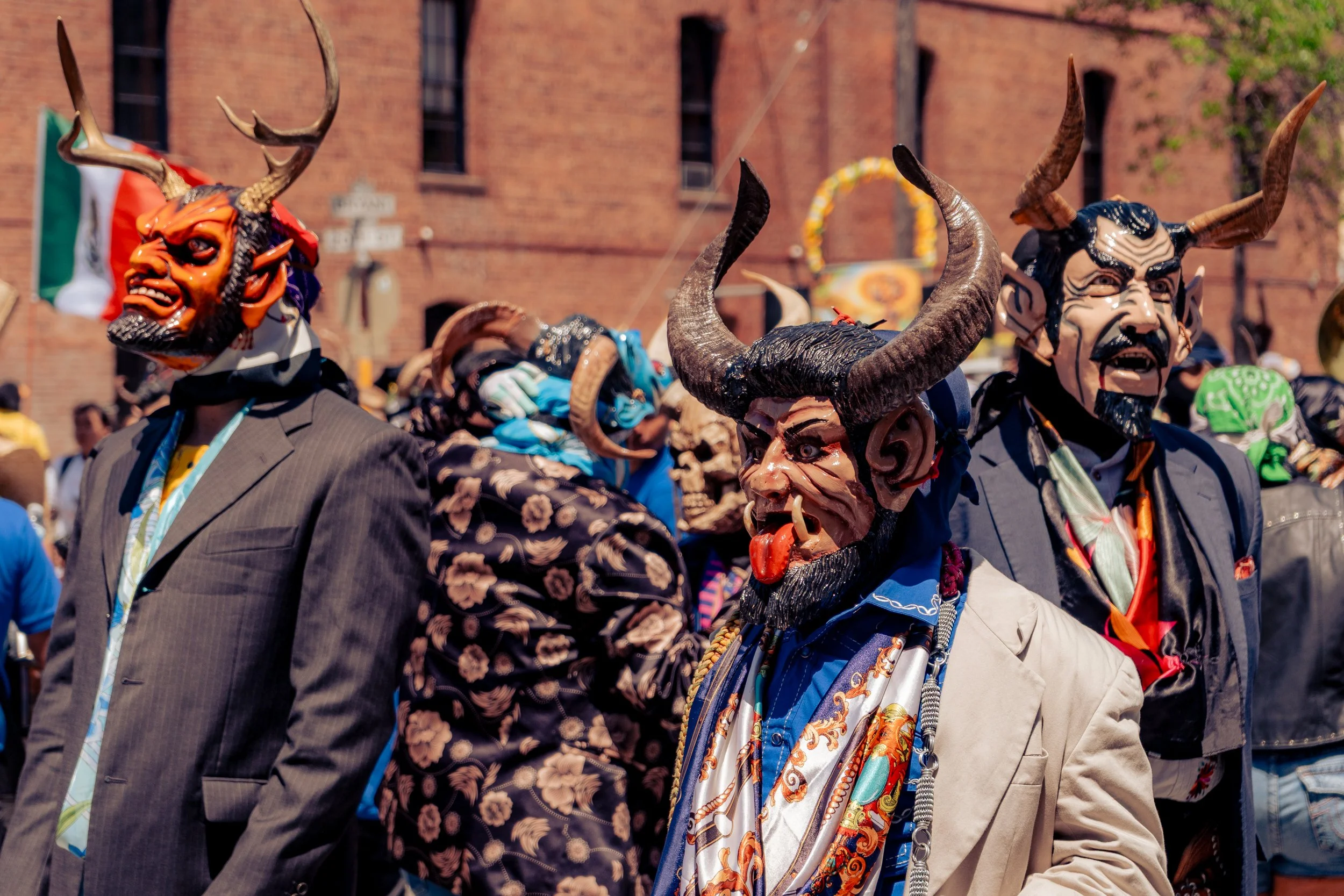 People wearing colorful masks and costumes during a cultural festival or celebration, with a brick building in the background.