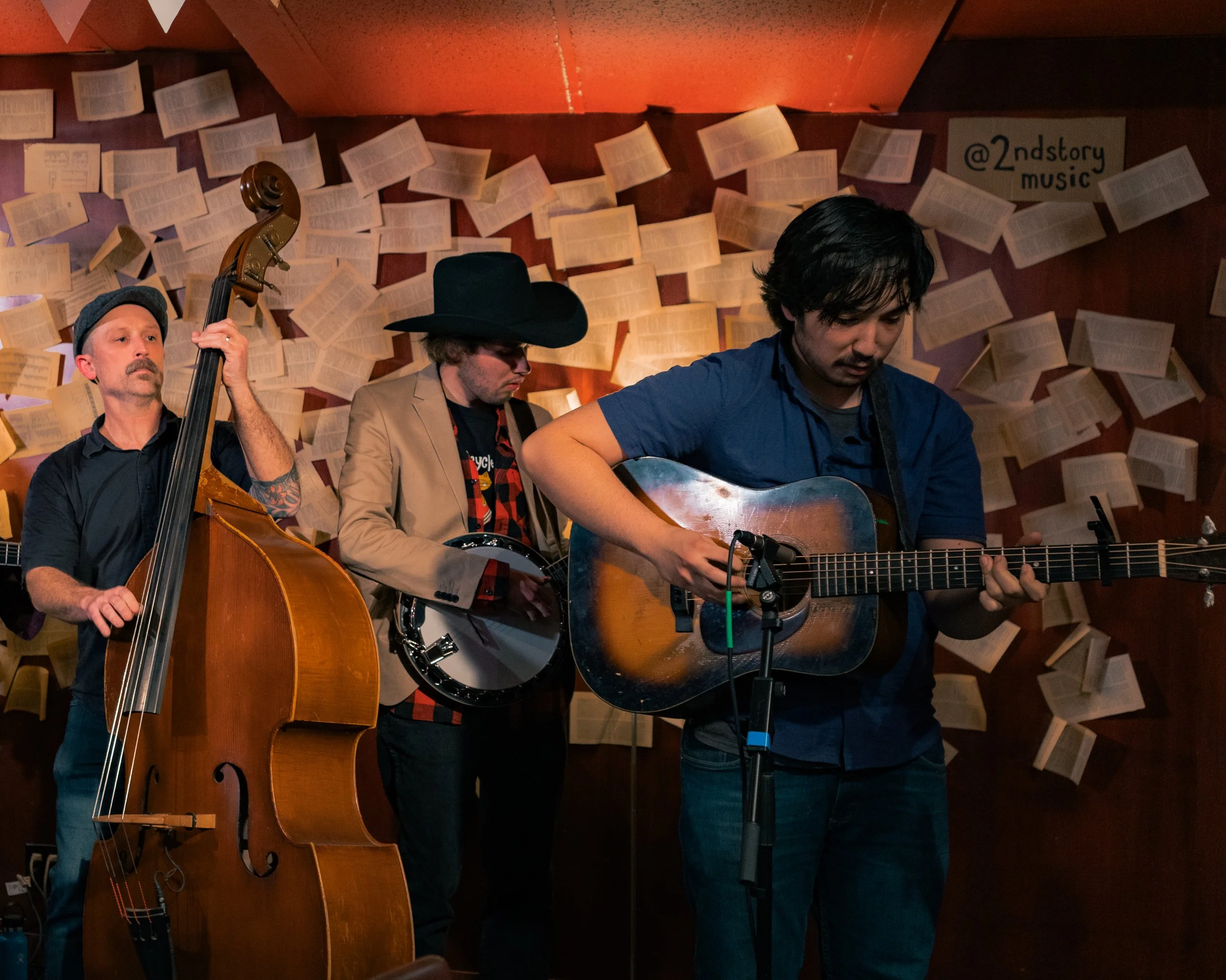 Three musicians playing instruments in a dimly lit venue with paper pages decorated on the wall behind them.