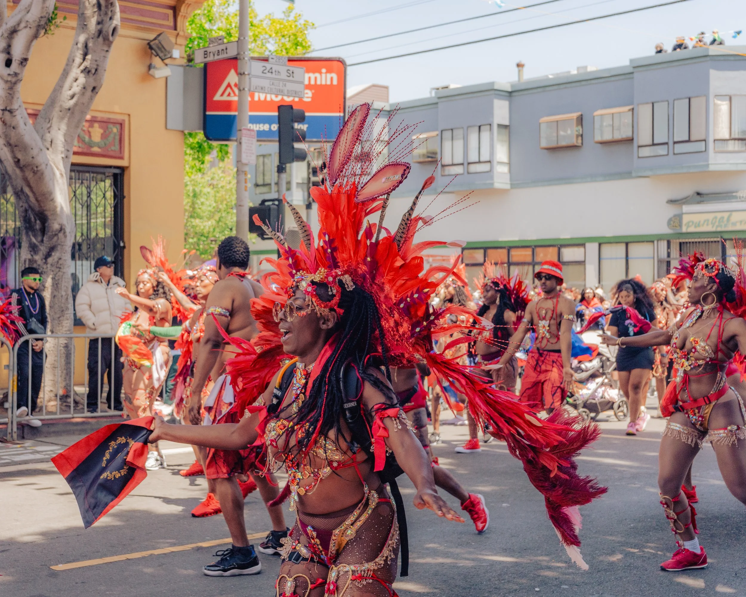 People in colorful costumes participating in a parade, with many wearing red feathered headdresses and jewelry, walking along a city street with onlookers and buildings in the background.