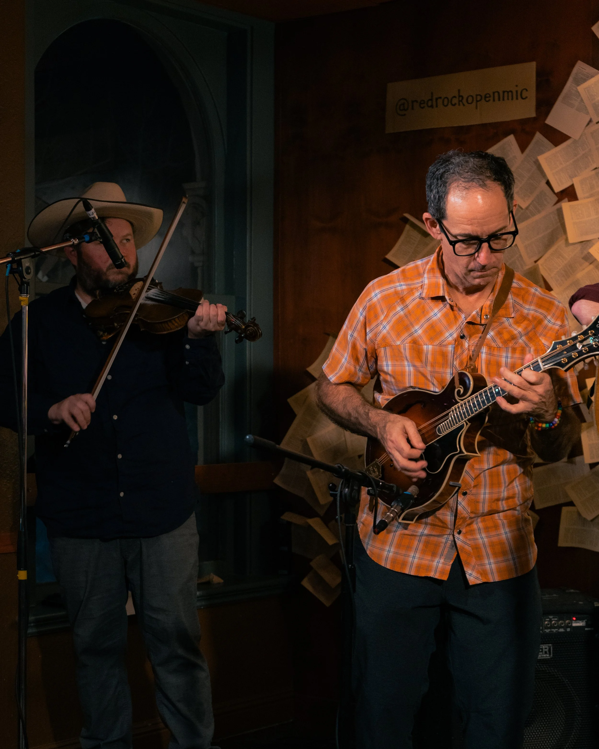 Two musicians performing indoors, one playing a violin and the other a mandolin, with sheet music and books pinned on the wall behind them.
