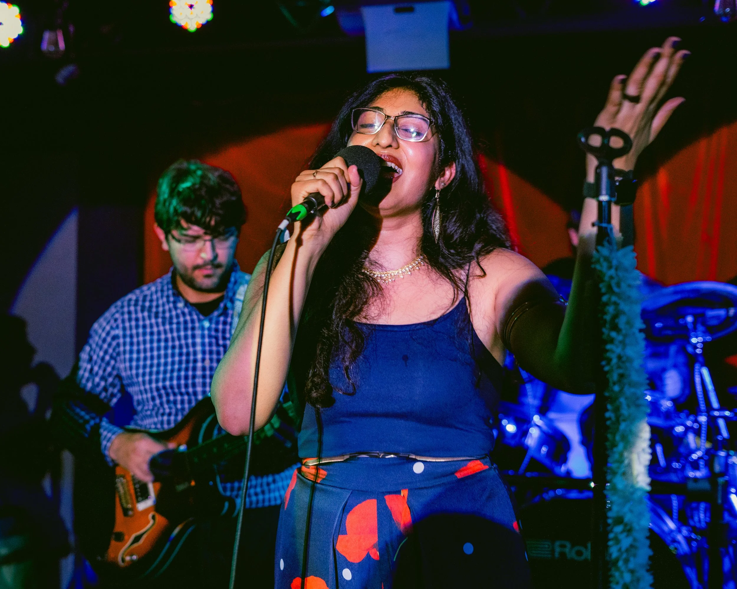 A woman singing into a microphone on stage with a guitarist in the background.