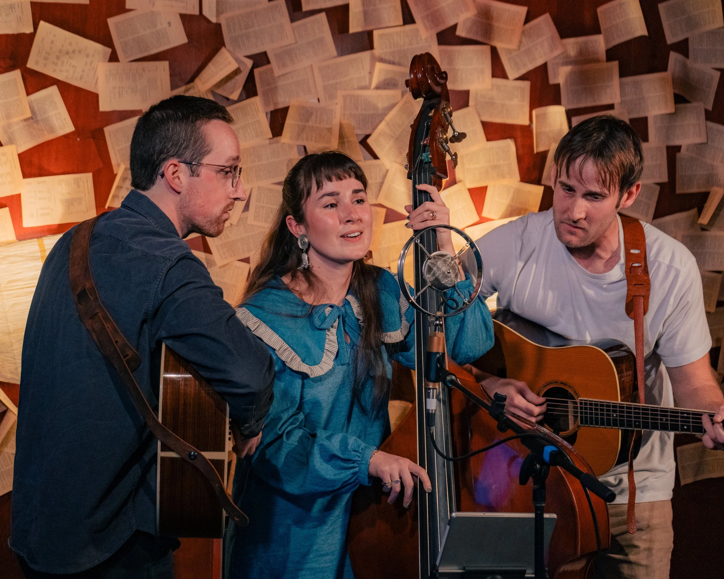 Three musicians performing with guitars and a microphone in a warmly lit room with numerous open books on the walls.