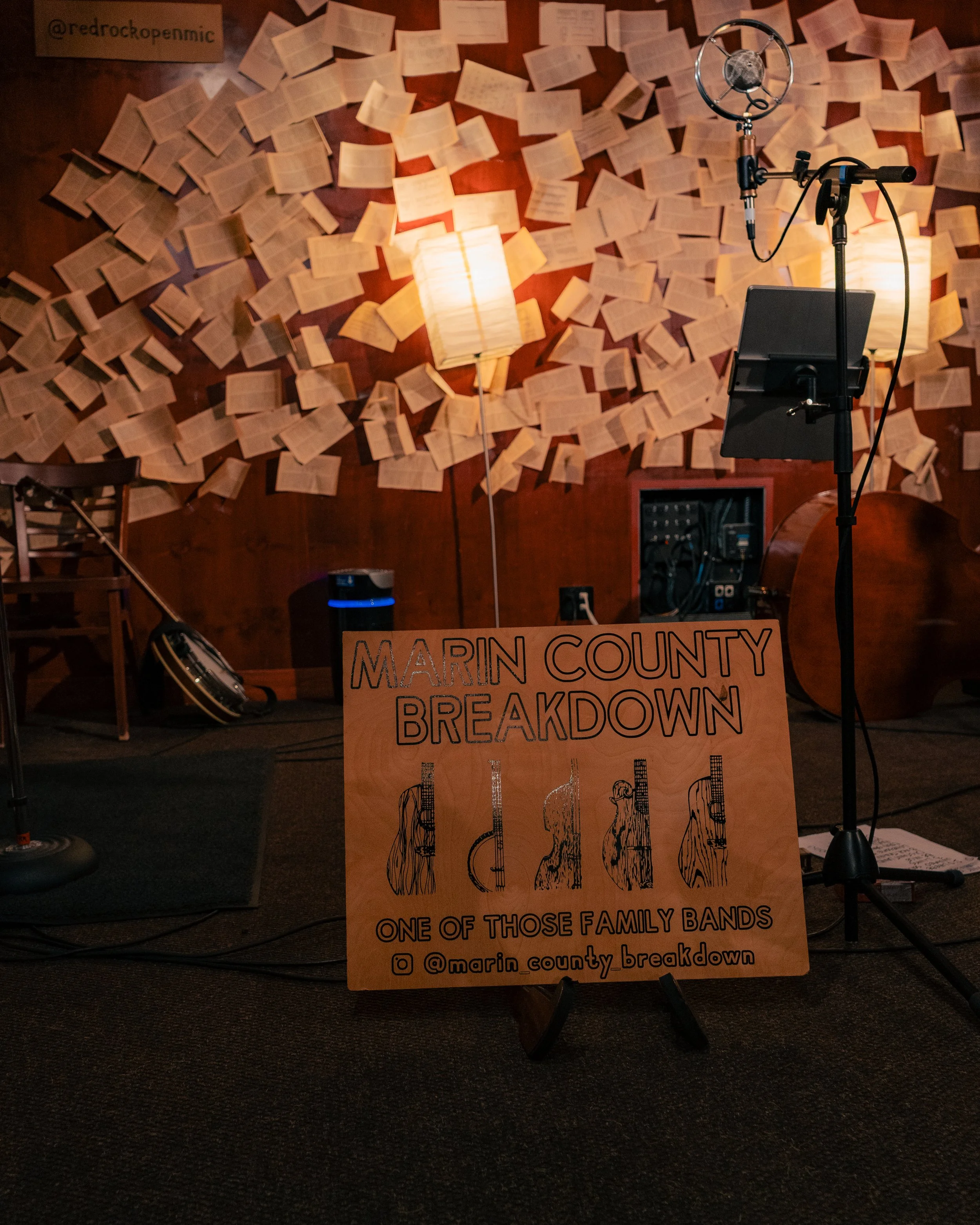 A cozy indoor music setup featuring a wooden sign that reads 'Marin County Breakdown,' with illustrations of different stringed instruments, and a social media handle '@marin_county_breakdown'. The background wall is decorated with open books or shee