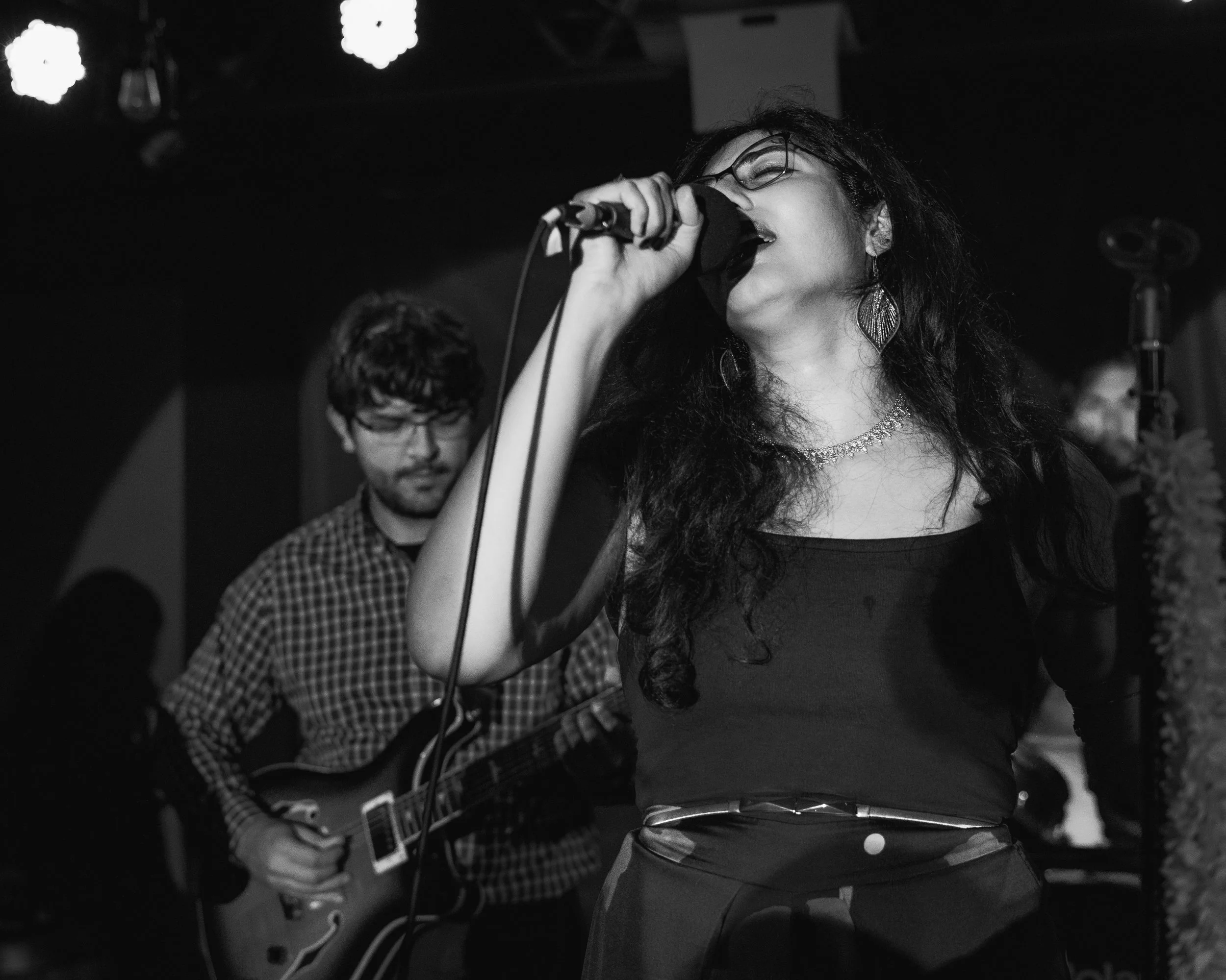A woman singing into a microphone with her eyes closed, wearing glasses, jewelry, and a dark outfit, while a man plays an electric guitar in the background in a dimly lit venue.