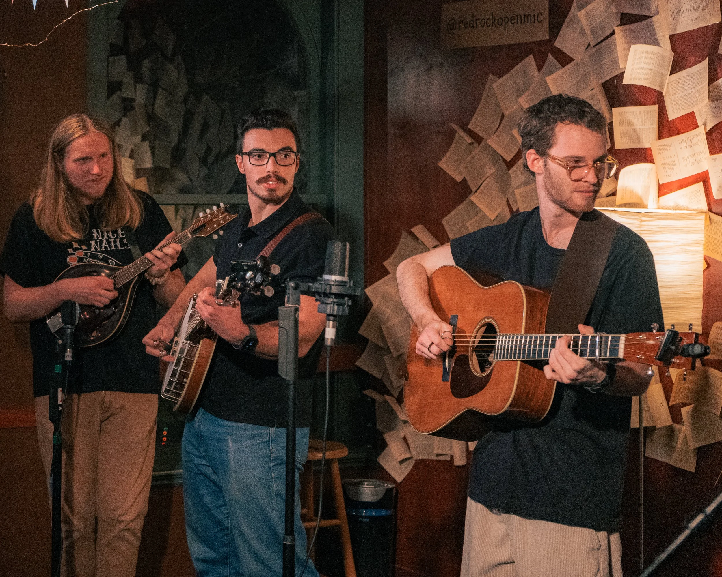 Three musicians performing indoors, two playing guitars and one playing a mandolin, with sheets of paper on the wall behind them.