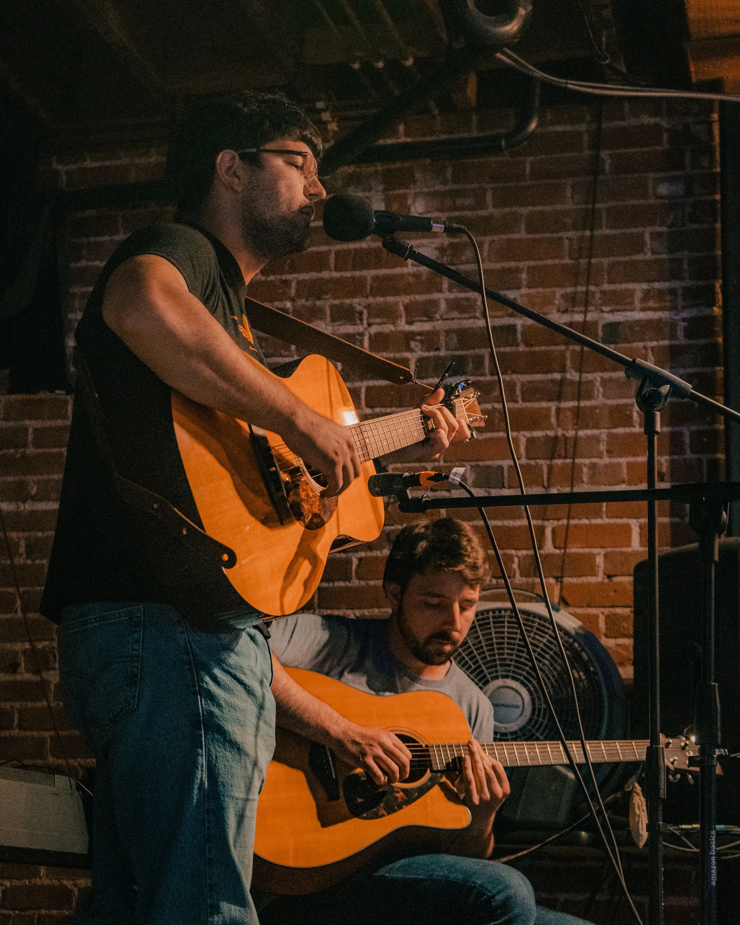 Two men playing acoustic guitars on a stage with a brick wall background, one singing into a microphone.