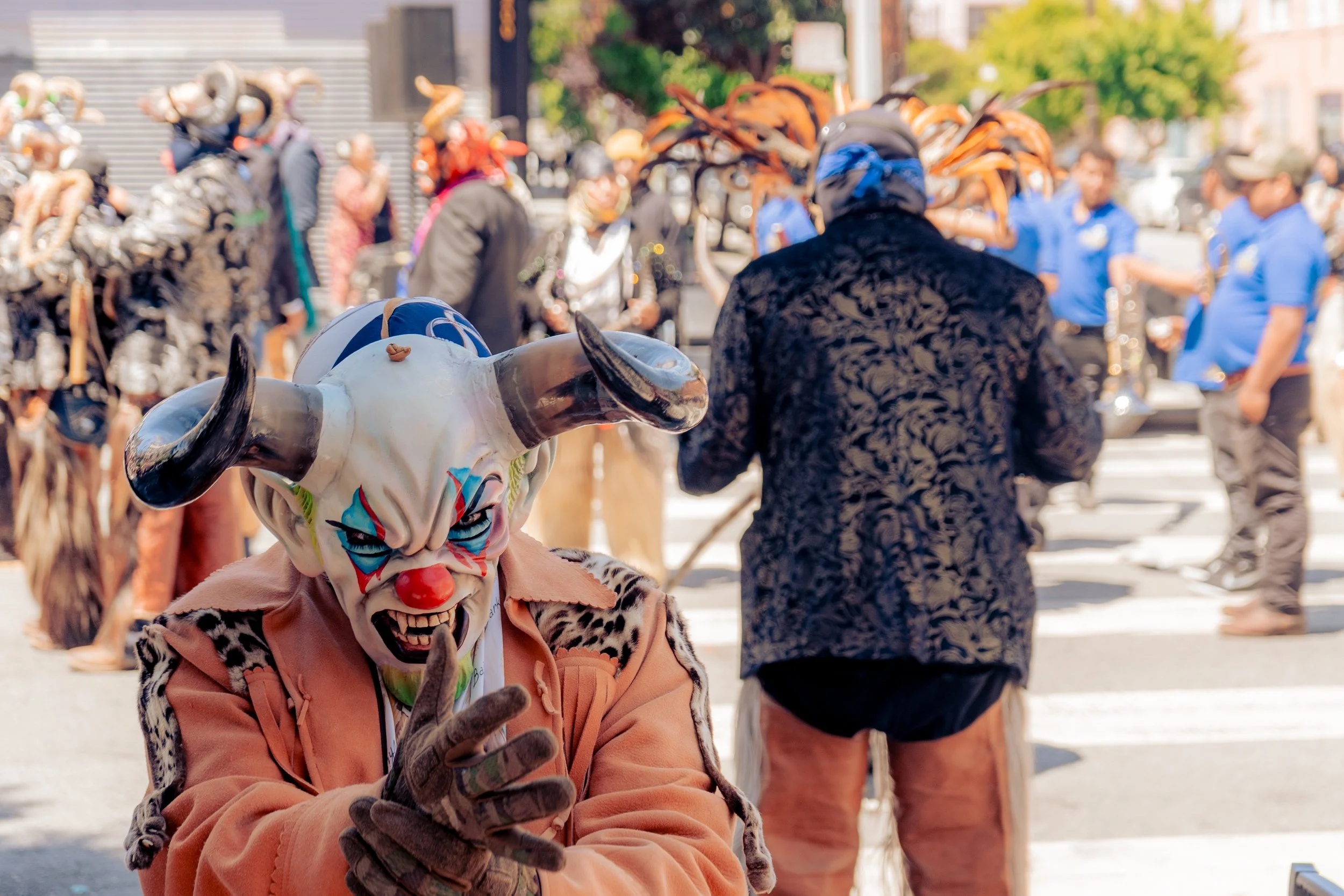 Person dressed as a clown with a sinister mask, horned face, and colorful paint, making a hand gesture during a street carnival with other costumed people and musicians in the background.
