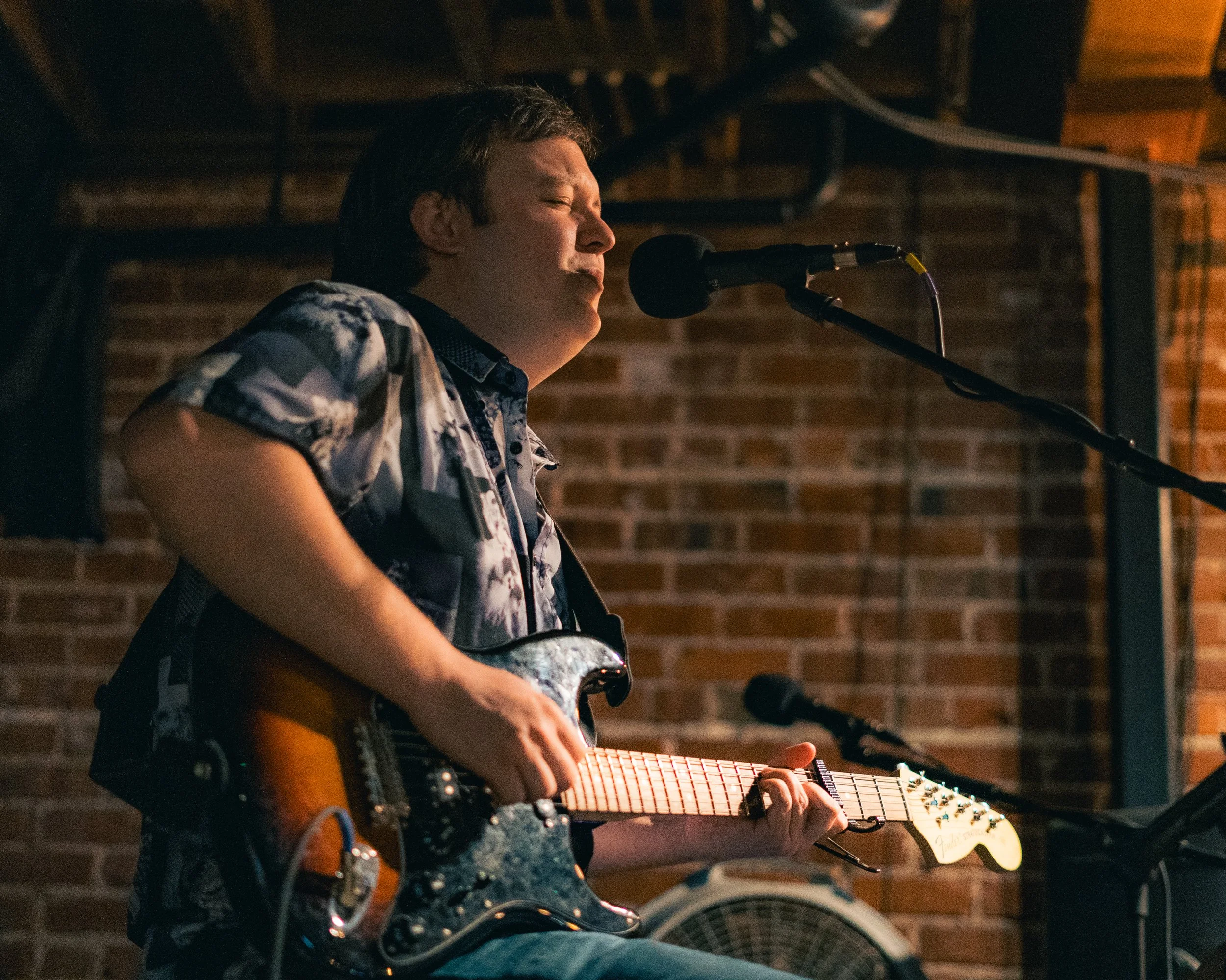 A young man performing live music with an electric guitar, singing into a microphone, in a dimly lit venue with brick walls.