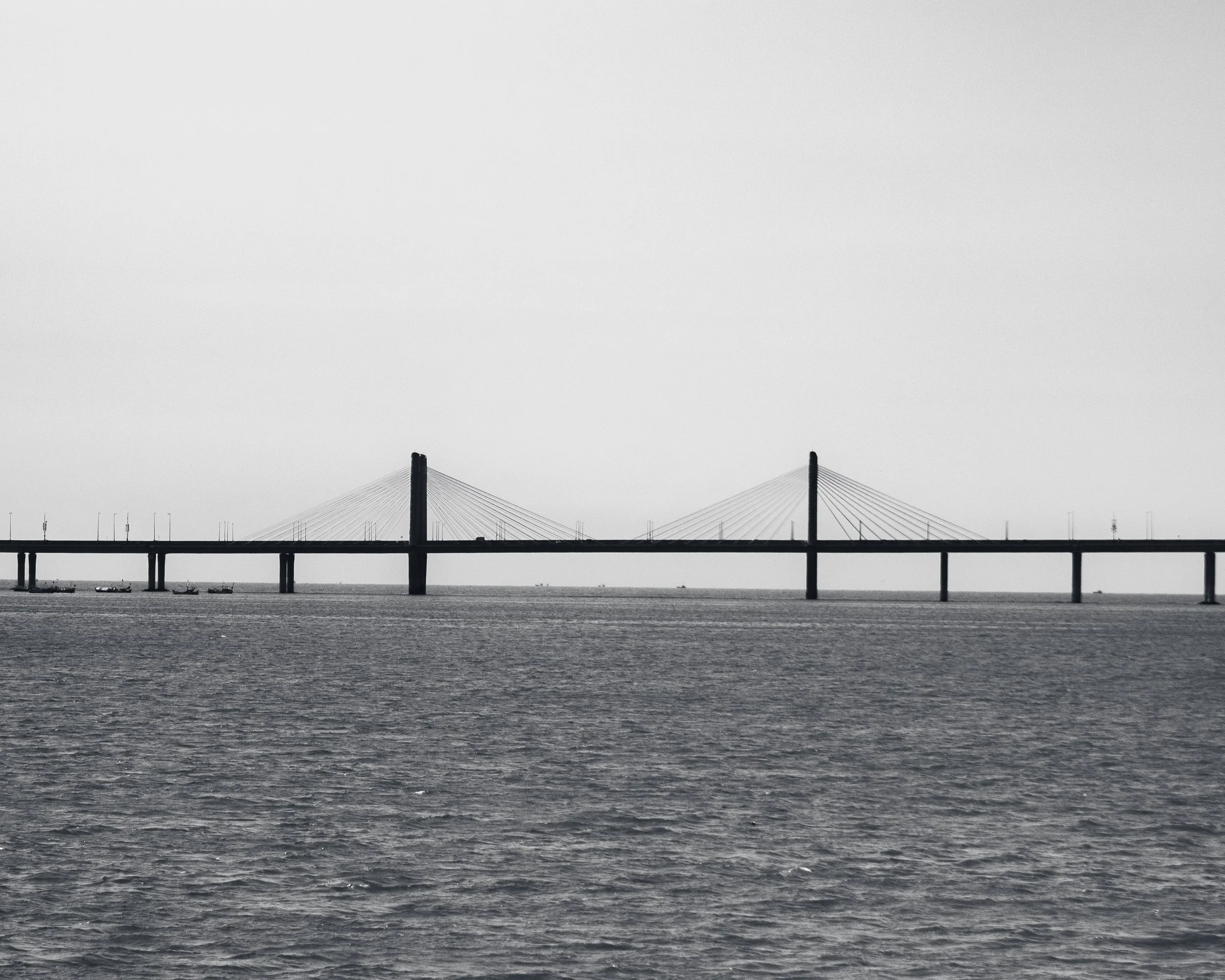 A black and white photo of a bridge over the water with two tall towers and cables, and several boats underneath.