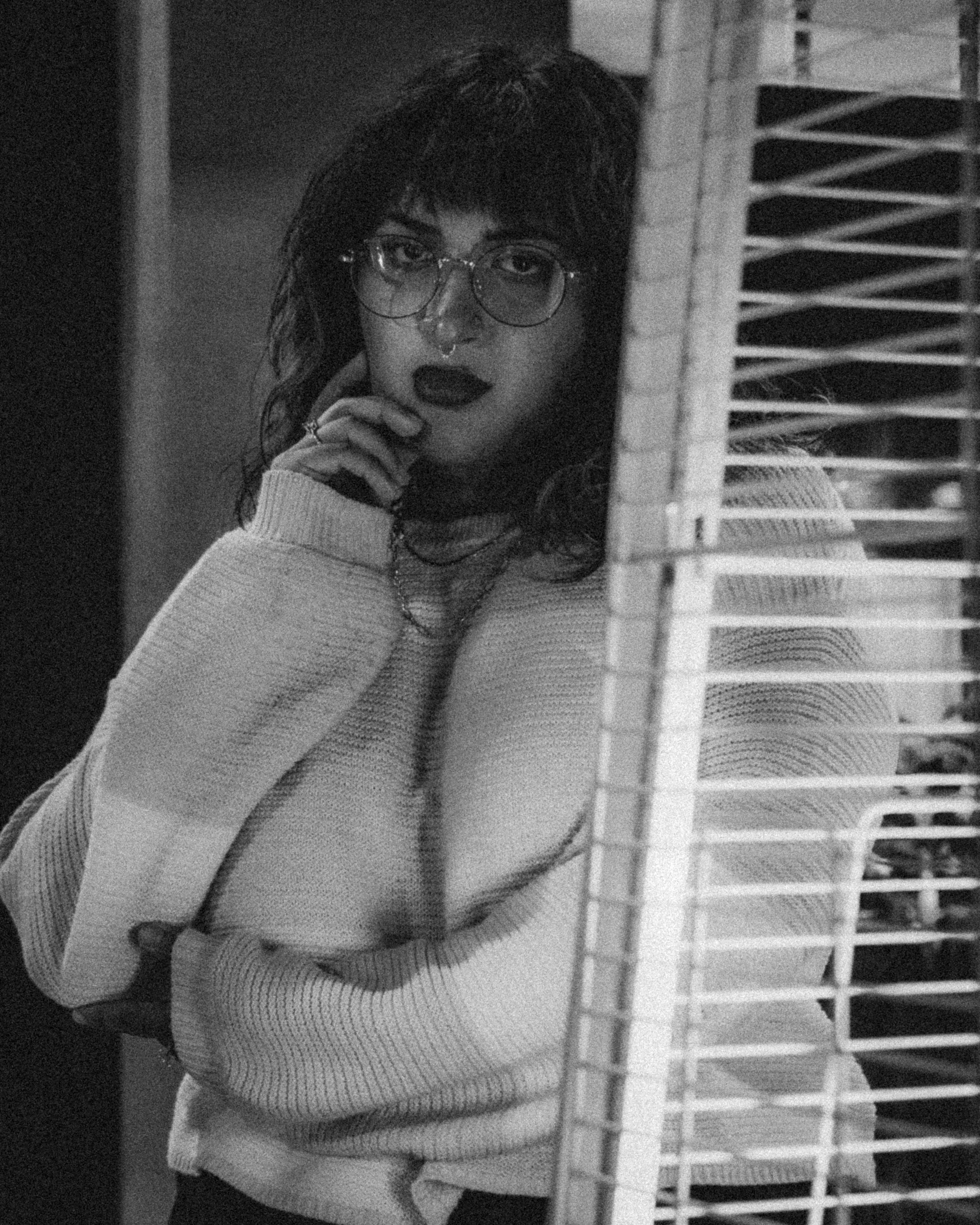 Black and white photo of a woman wearing glasses, a septum ring, dark lipstick, and a light-colored sweater, looking at the camera with her hand near her face, partially obscured by window blinds.