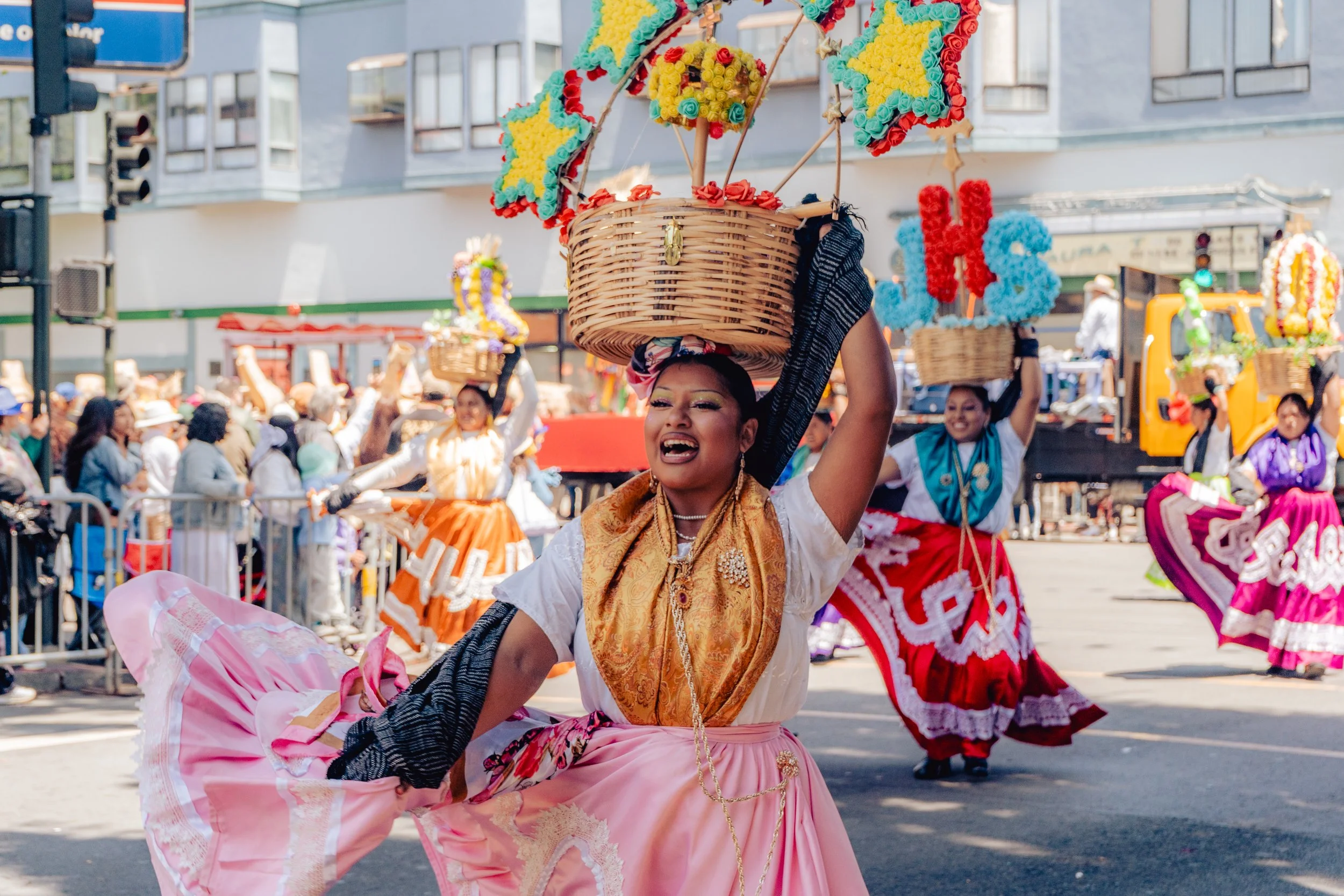 Women dancing in traditional colorful Mexican dresses and skirts during a parade, carrying baskets decorated with flowers on their heads. Crowd of spectators in the background.