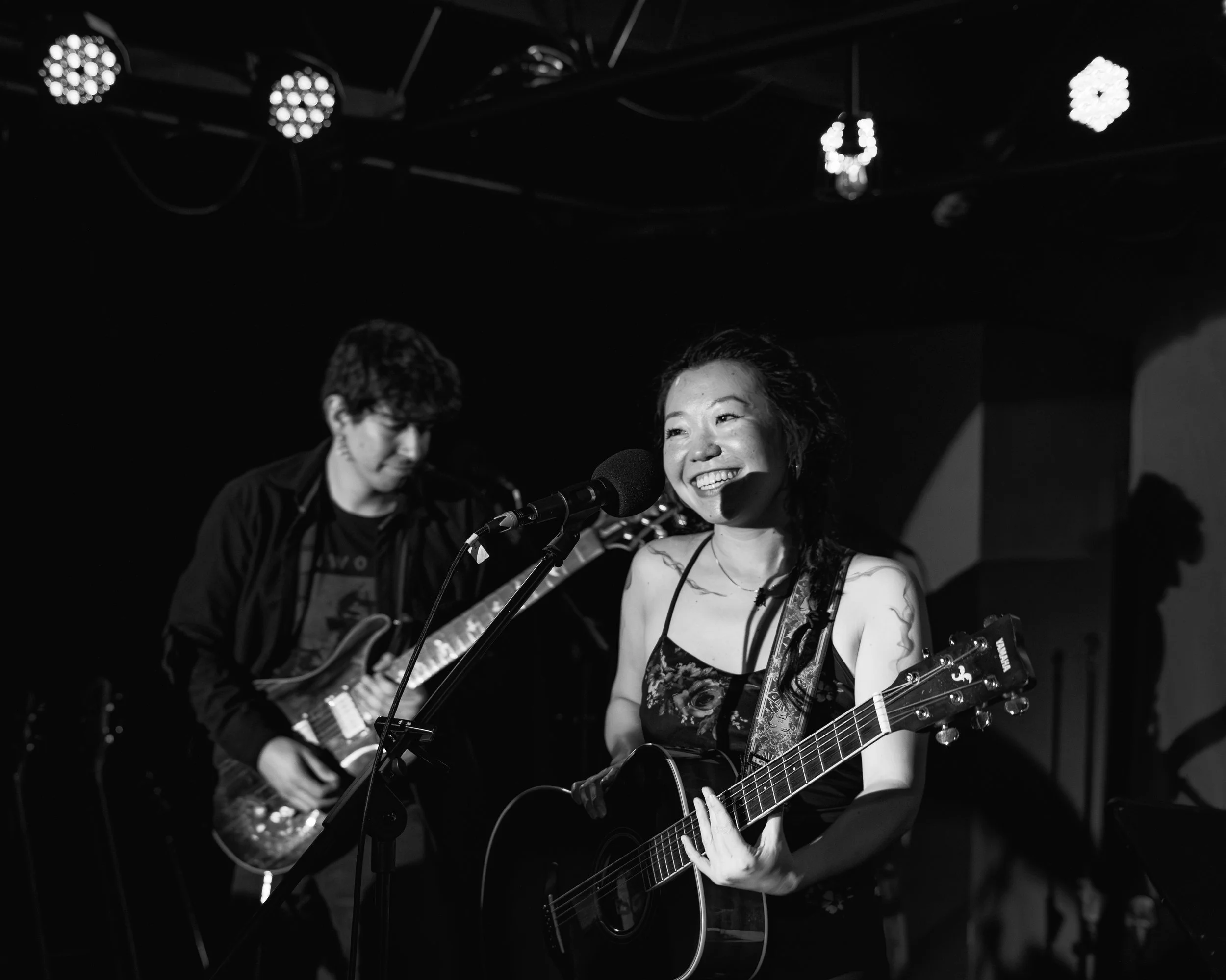 A woman playing an acoustic guitar and singing into a microphone, smiling, with a man in the background playing an electric guitar, on a stage with overhead lights.