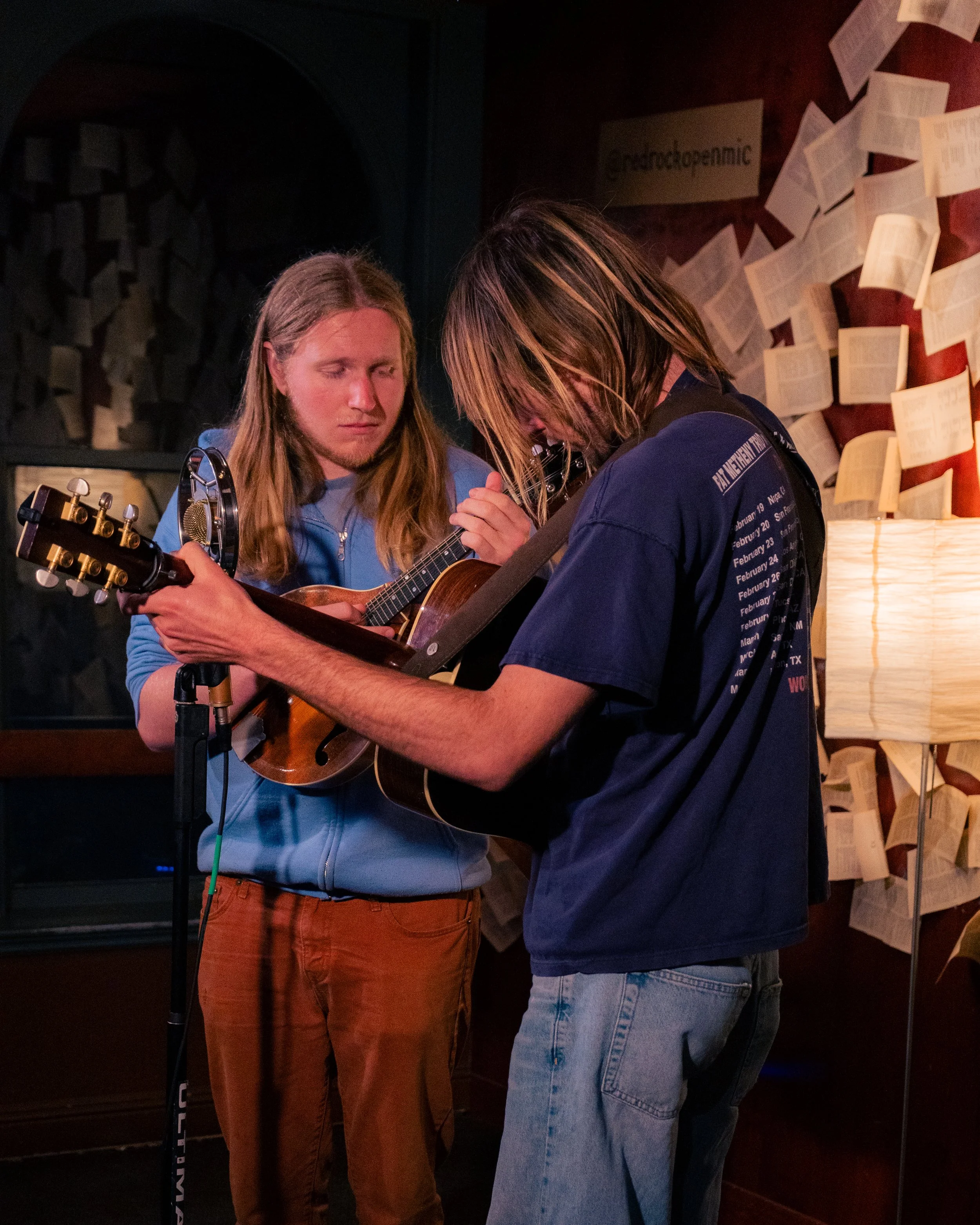 Two young men playing acoustic guitar together in a dimly lit room decorated with papers on the wall.