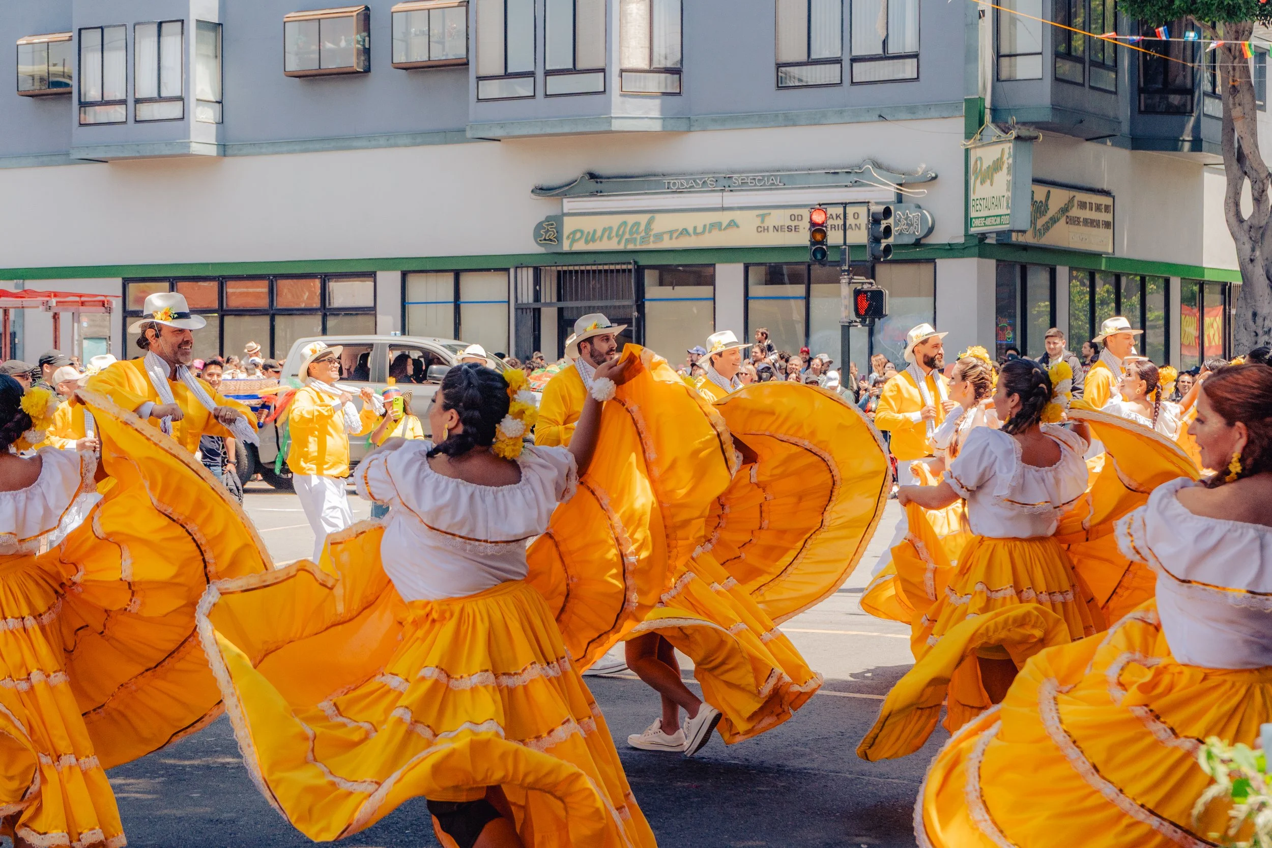 A lively street parade featuring women in white blouses and vibrant yellow skirts, and men in yellow jackets and hats, dancing and celebrating in front of a store with a crowd of spectators watching.