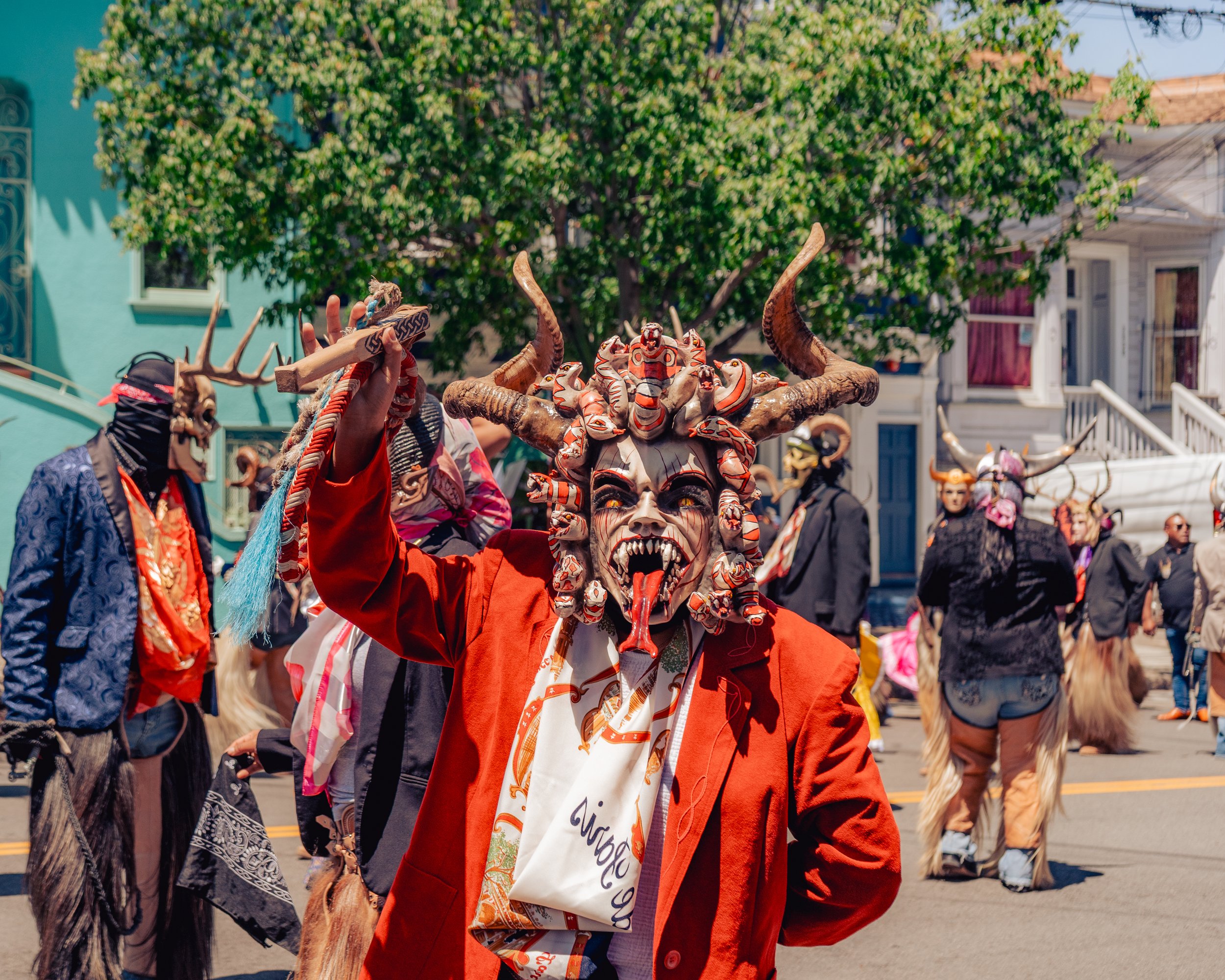 People in costumes participating in a cultural parade, wearing masks with horns and animal features, in an outdoor street setting with colorful buildings and trees in the background.