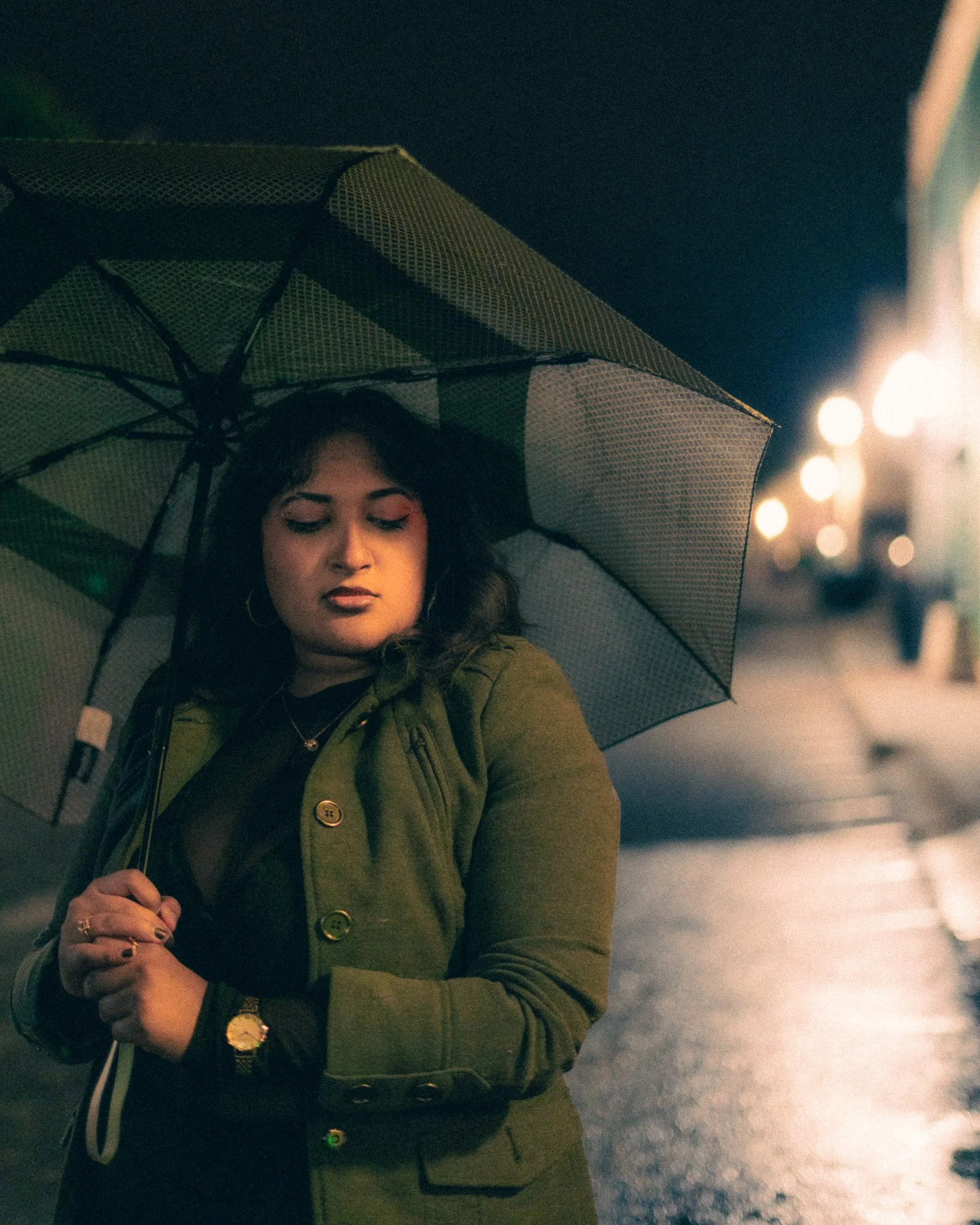 A woman with dark hair stands under a black and white checkered umbrella on a nighttime street, looking down with a contemplative expression. The street is wet and illuminated by blurred street lights.