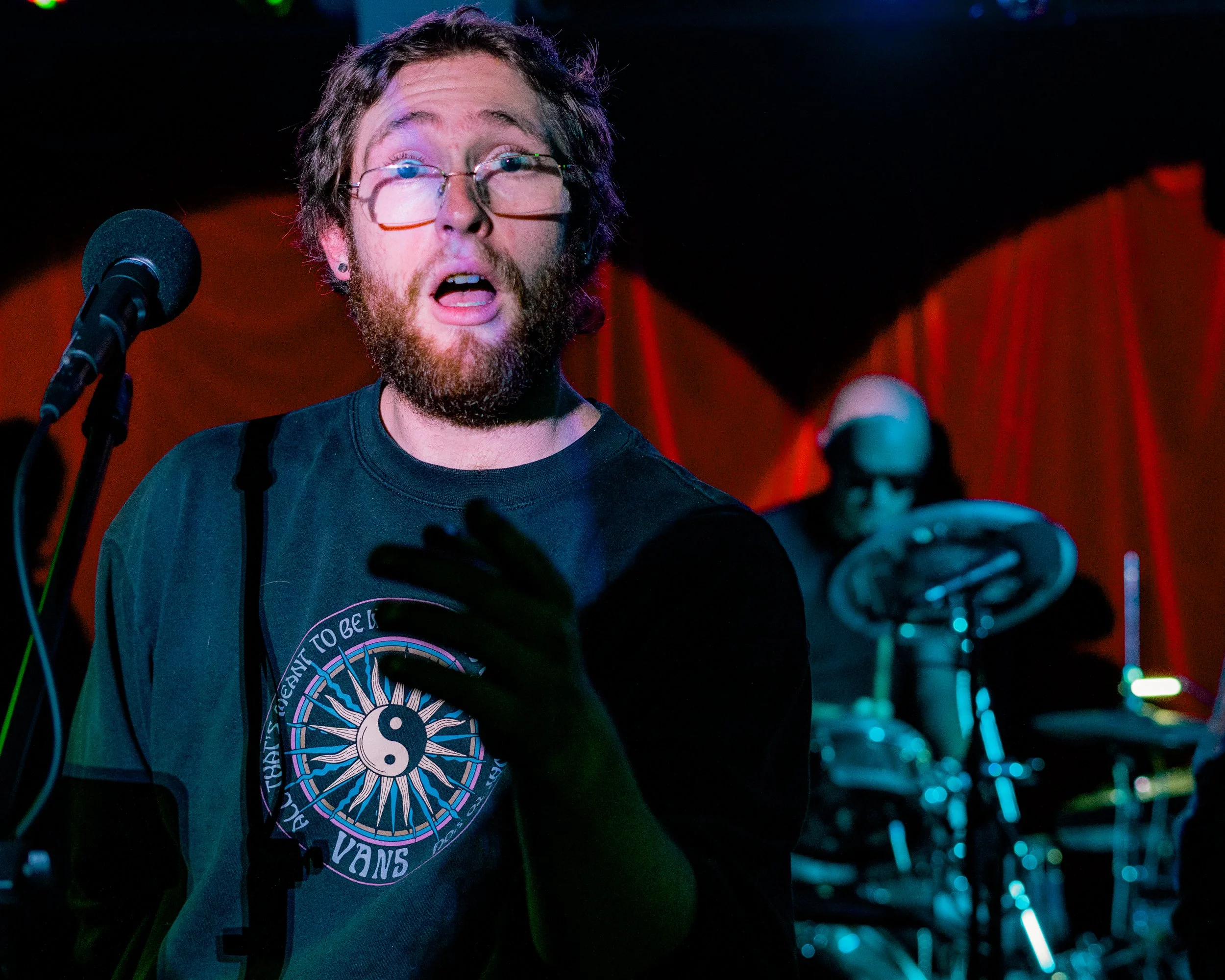 A man with glasses and a beard singing into a microphone on stage, with a drummer in the background. The stage has red curtains.