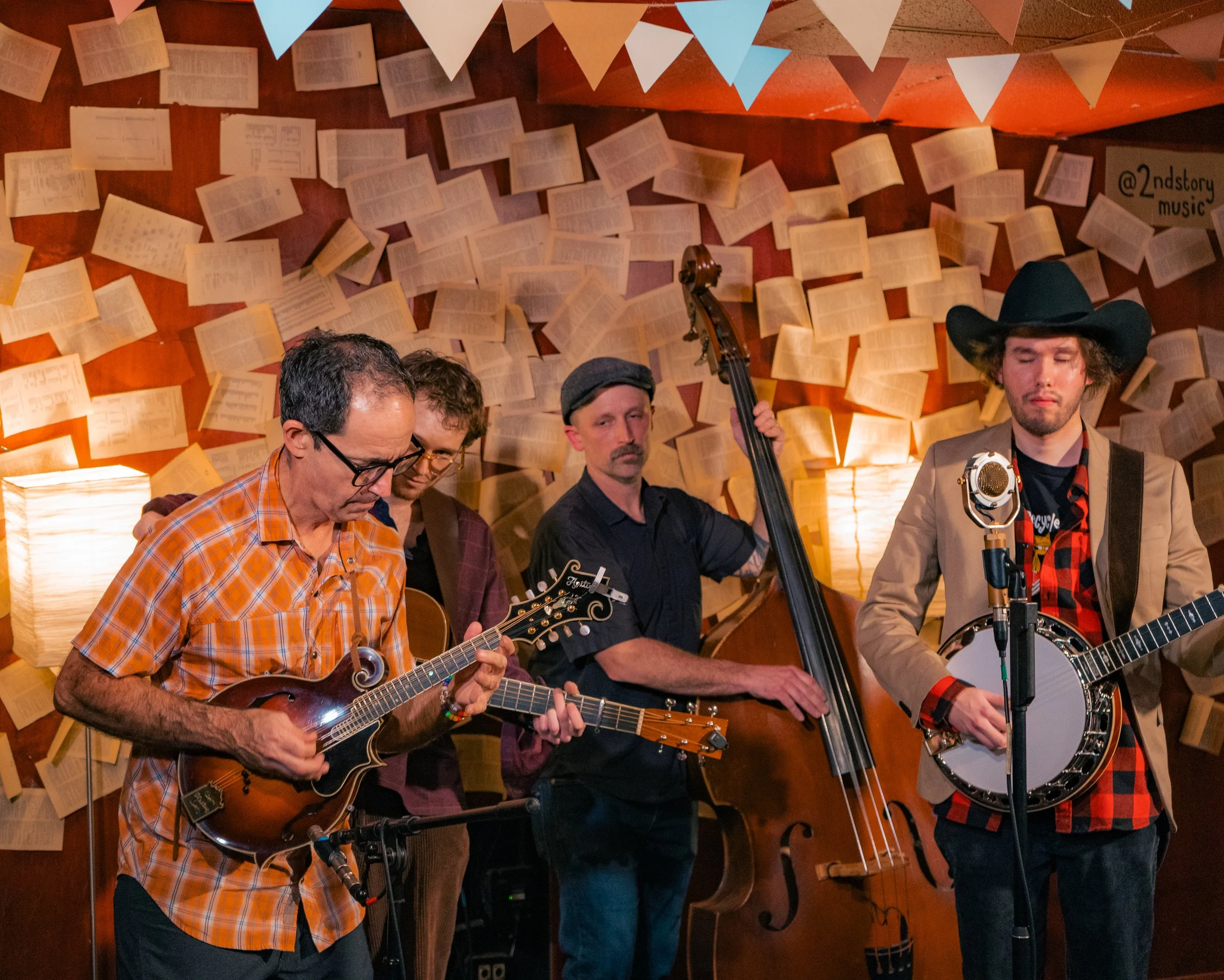 Four musicians performing with acoustic and electric guitars and a double bass in a room decorated with open books and paper banners.