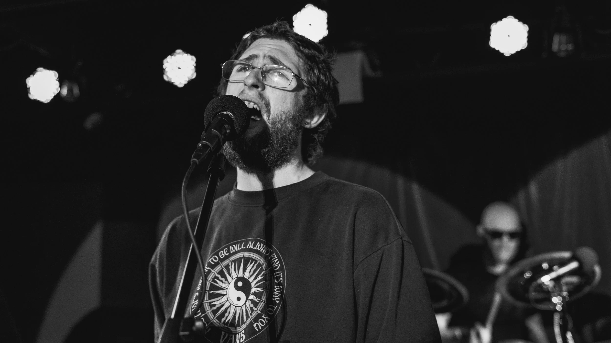 A man with glasses and a beard sings into a microphone on stage, with a drummer in sunglasses playing in the background, in black and white.