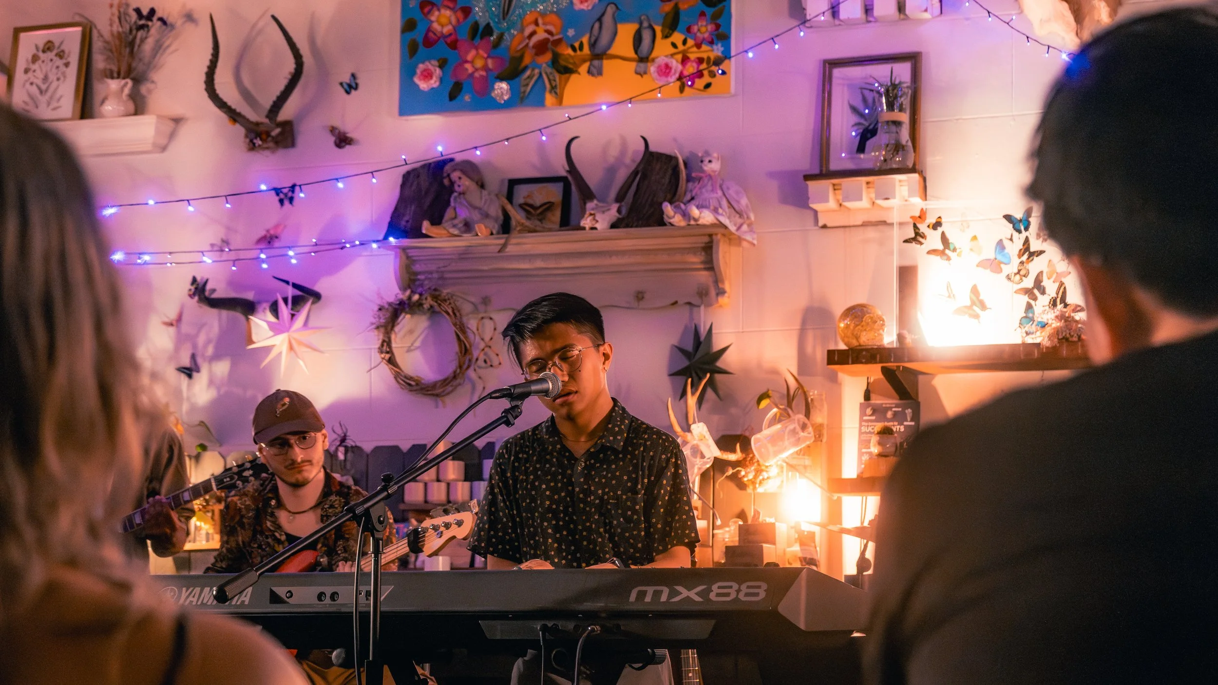 A group of musicians performing in a cozy, decorated room with string lights, artwork, and decorative objects. One musician is singing into a microphone while playing a keyboard, with another playing guitar in the background. The room has warm lighti