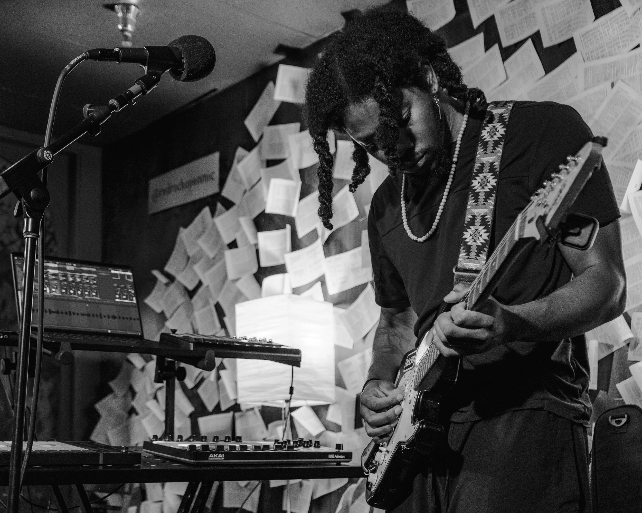 A person playing electric guitar in a music studio with a laptop and a mixing console, surrounded by numerous paper pages on the wall.