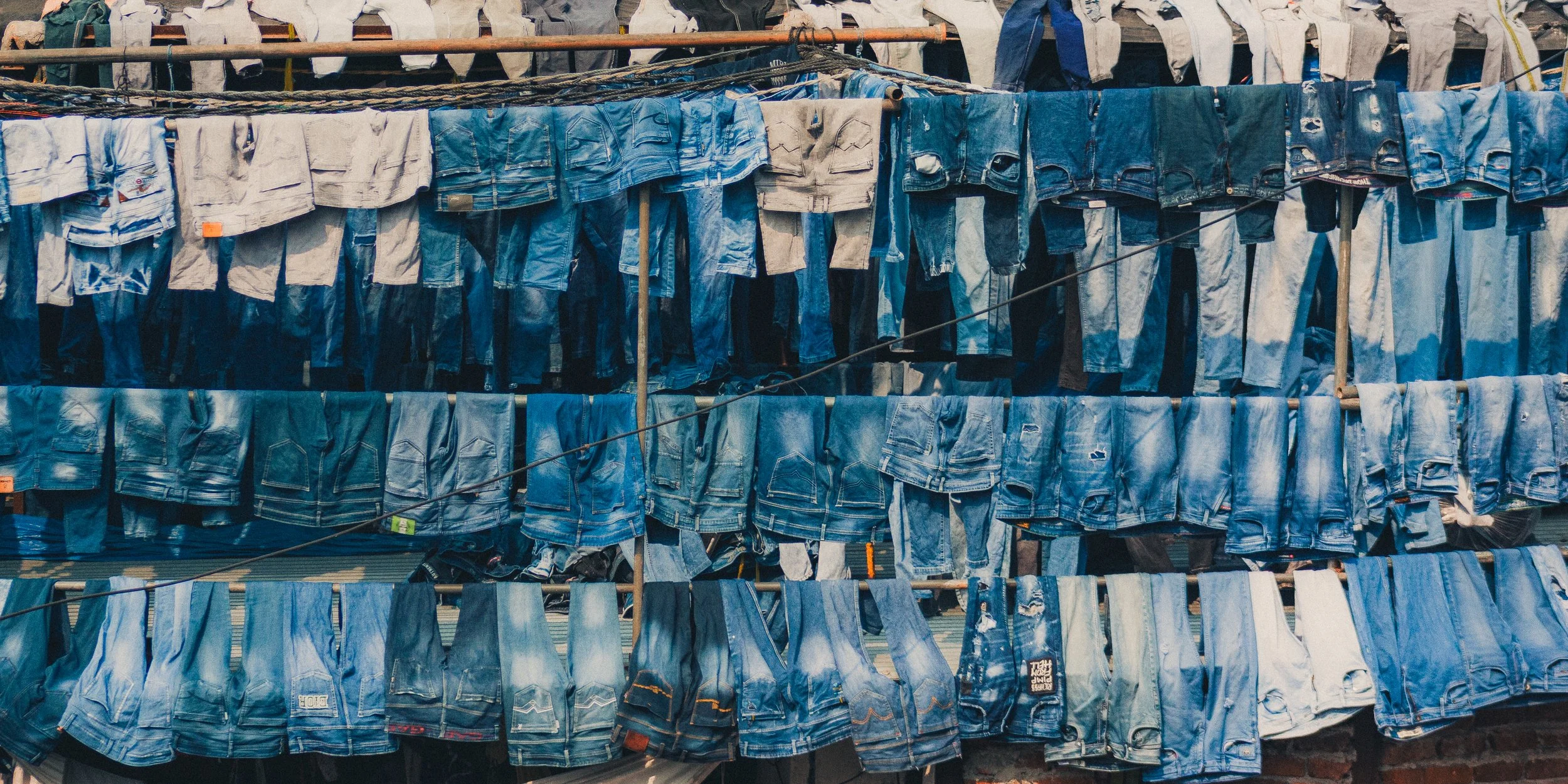 Multiple rows of denim jeans hanging on clotheslines in an outdoor market, with various shades of blue and some ripped or distressed jeans.