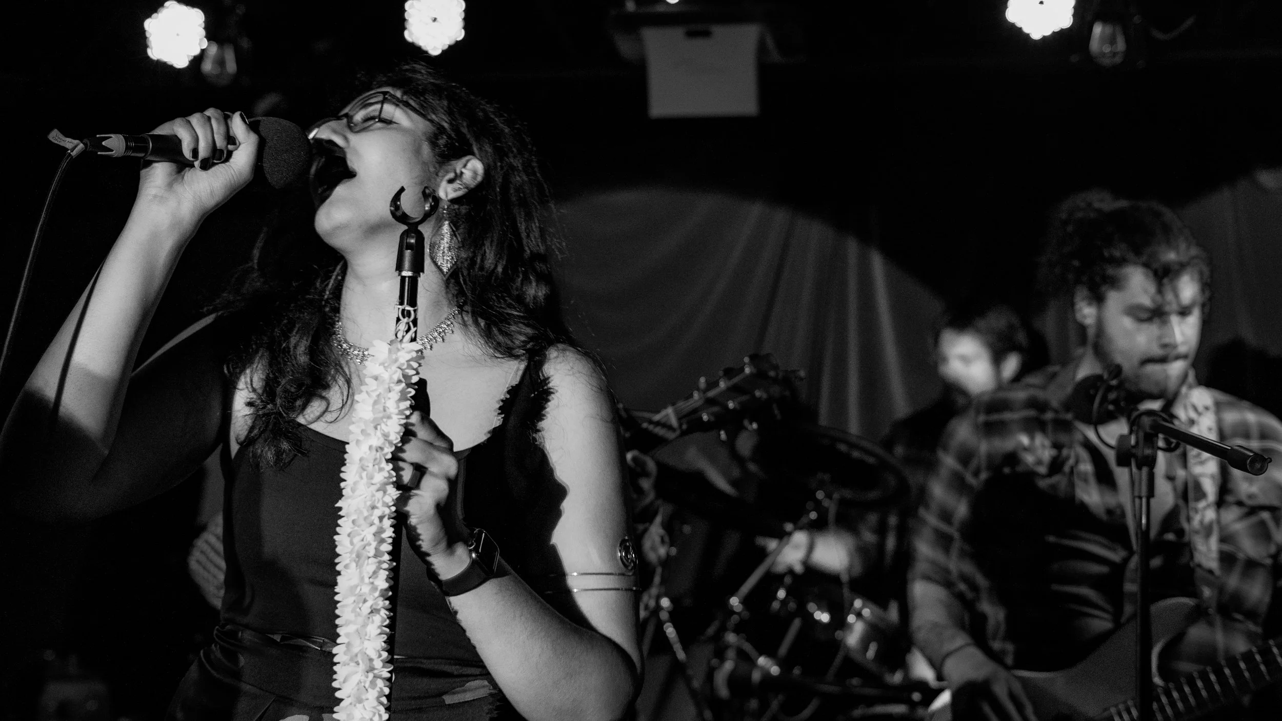 A female singer passionately performing on stage with her eyes closed, holding a microphone in one hand and a microphone stand with leis hanging from her neck. Two male guitar players are visible in the background, one on the left and one on the righ