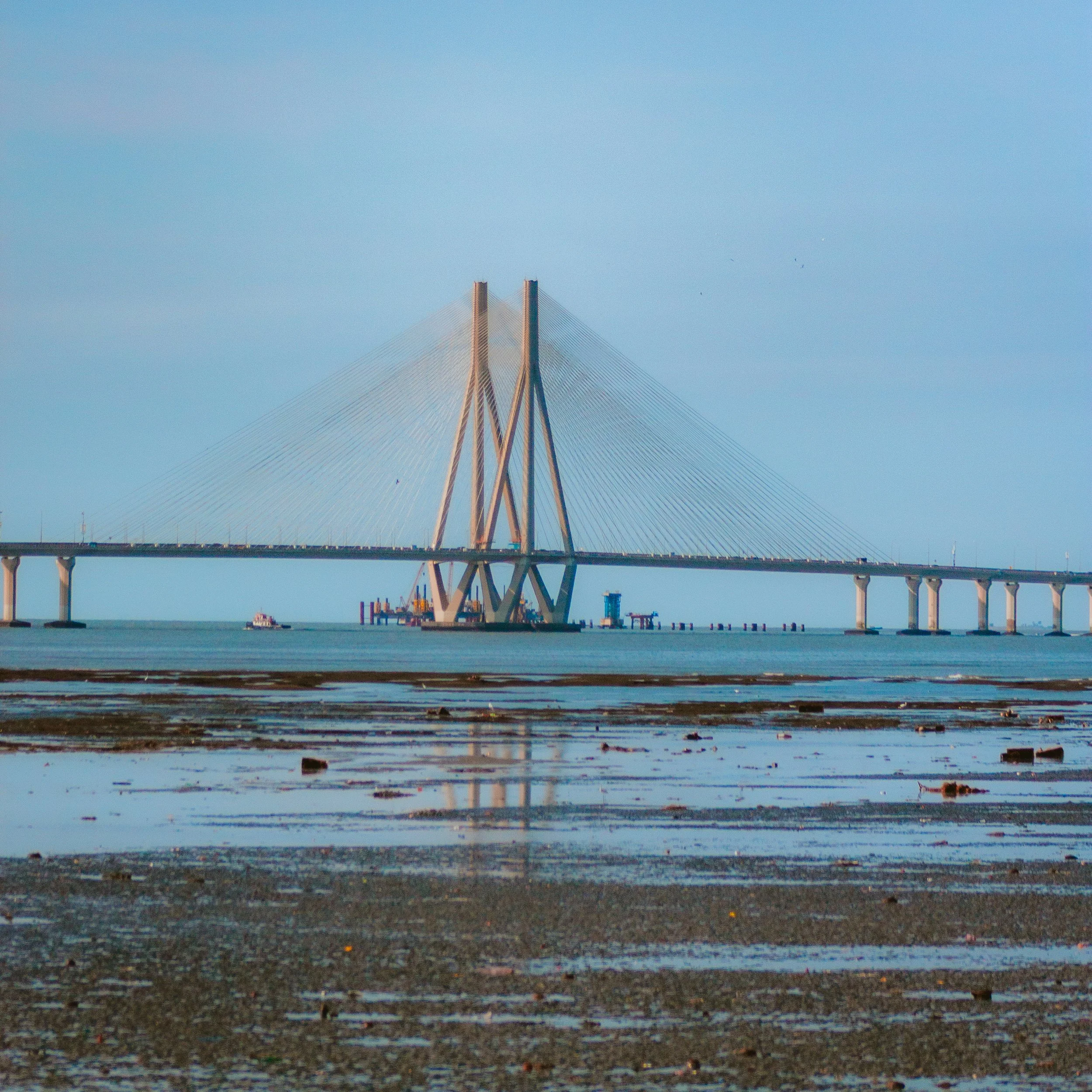 A large cable-stayed bridge under construction over water, with a sandy and rocky shore in the foreground and a clear sky in the background.