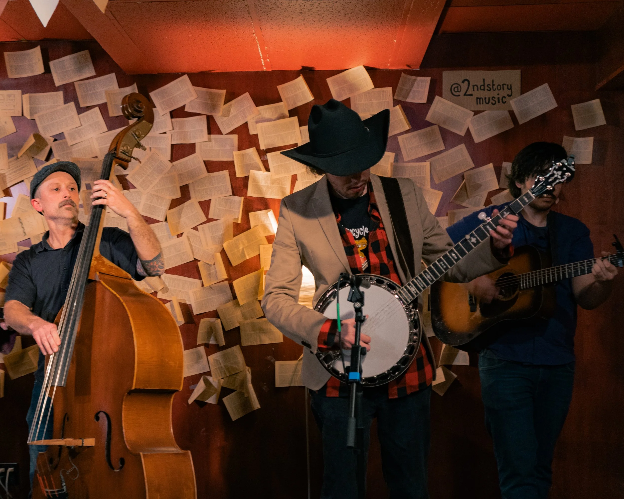 Three musicians performing on stage, playing upright bass, banjo, and acoustic guitar, with open books on the wall behind them and a sign that reads '@2ndstory music'.