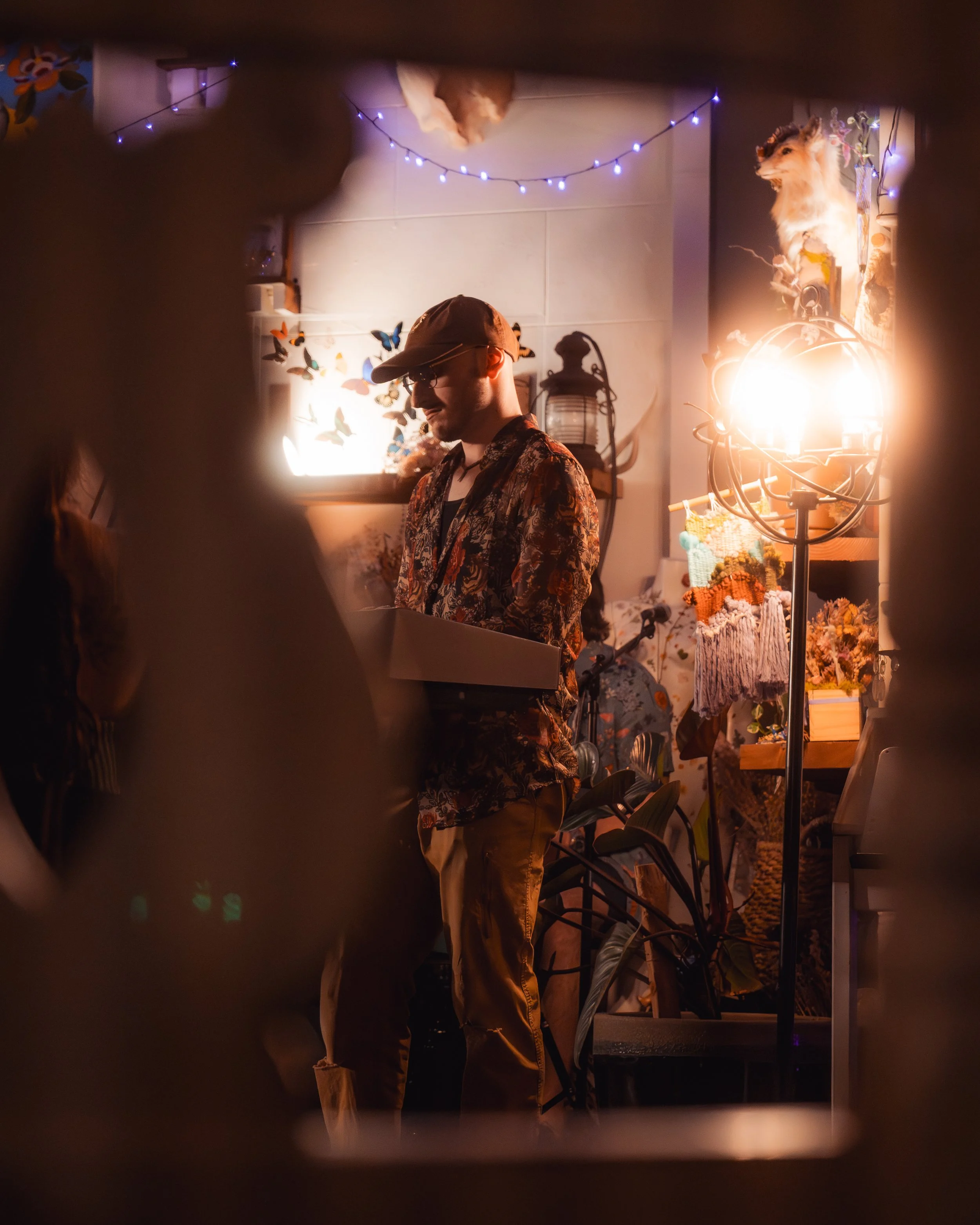 A man with a hat and sunglasses holding a cake box, standing in a cozy, decorated room with string lights, butterfly wall art, and a portrait of a lion.
