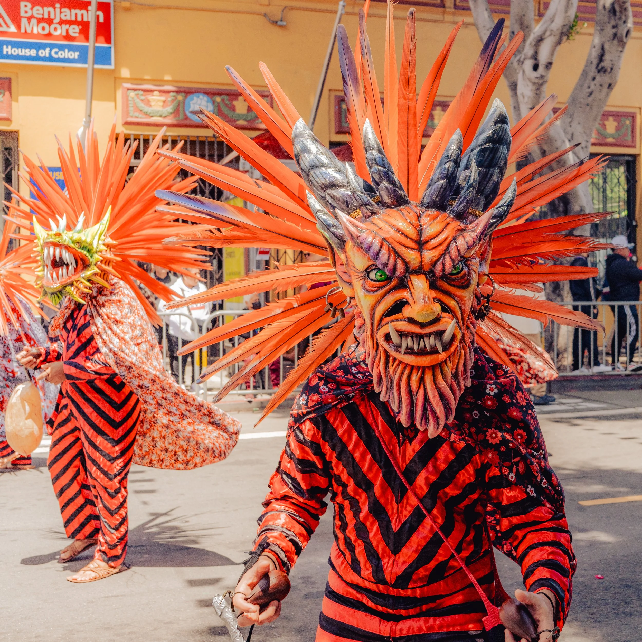 People participating in a parade wearing colorful, elaborate costume masks and outfits, featuring large masks resembling mythical creatures with fierce faces and feathered headdresses.