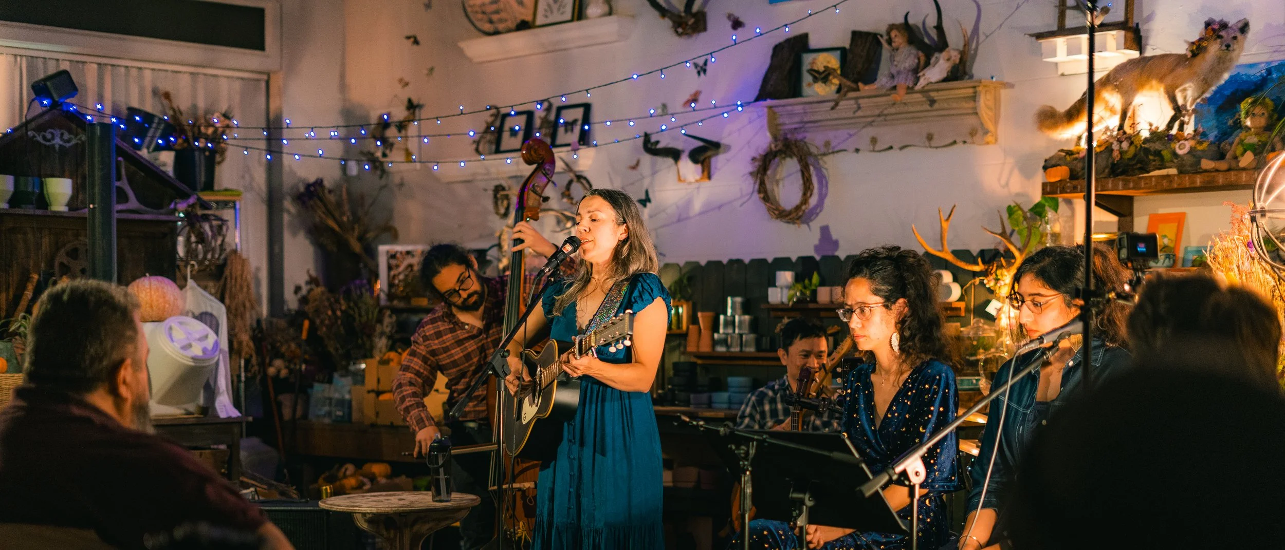 A woman with long hair sings and plays guitar and bass during a live music performance in a cozy, decorated indoor space, with several other musicians and audience members present.