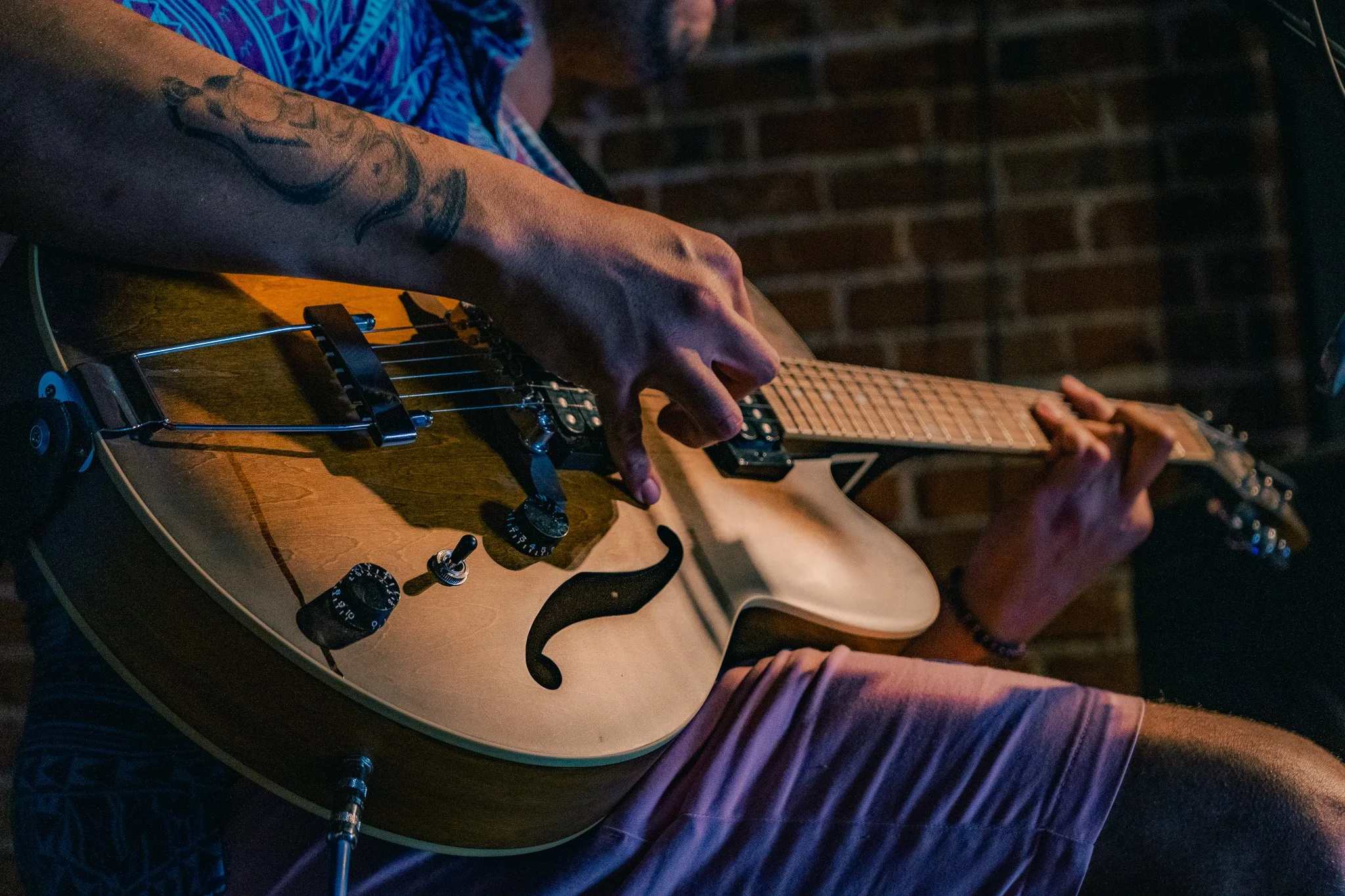 A person playing an electric guitar with a tattooed arm, seated against a brick wall.