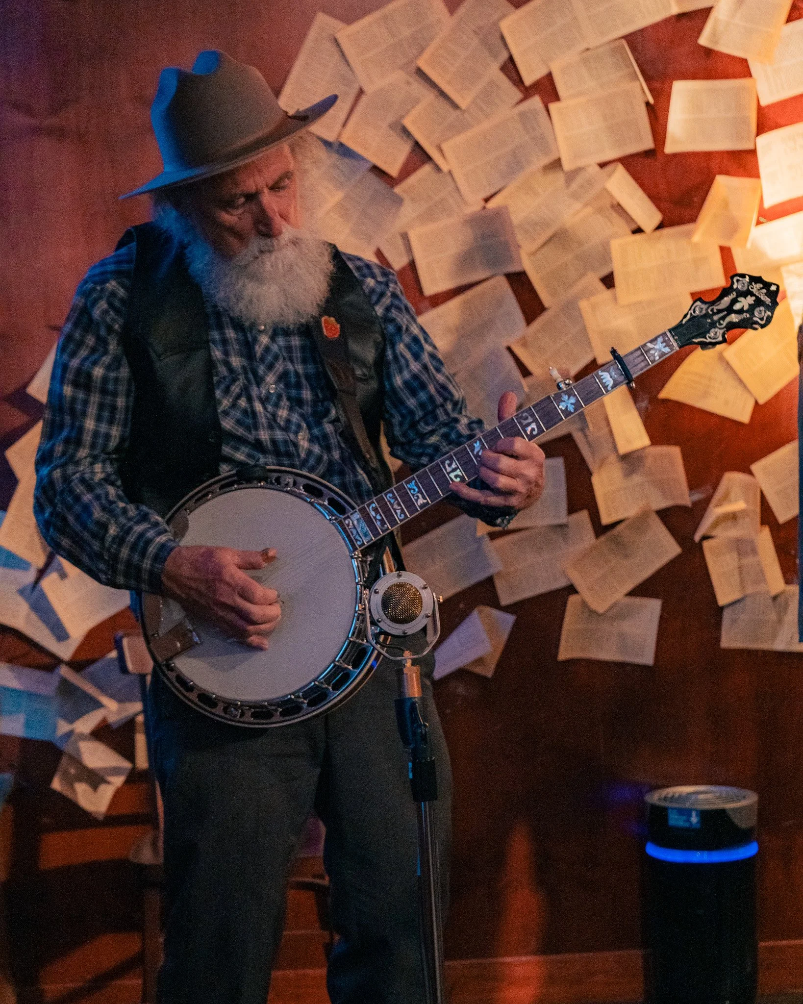 An older man with a white beard wearing a cowboy hat and checkered shirt, playing a banjo in a room with walls covered in open books.
