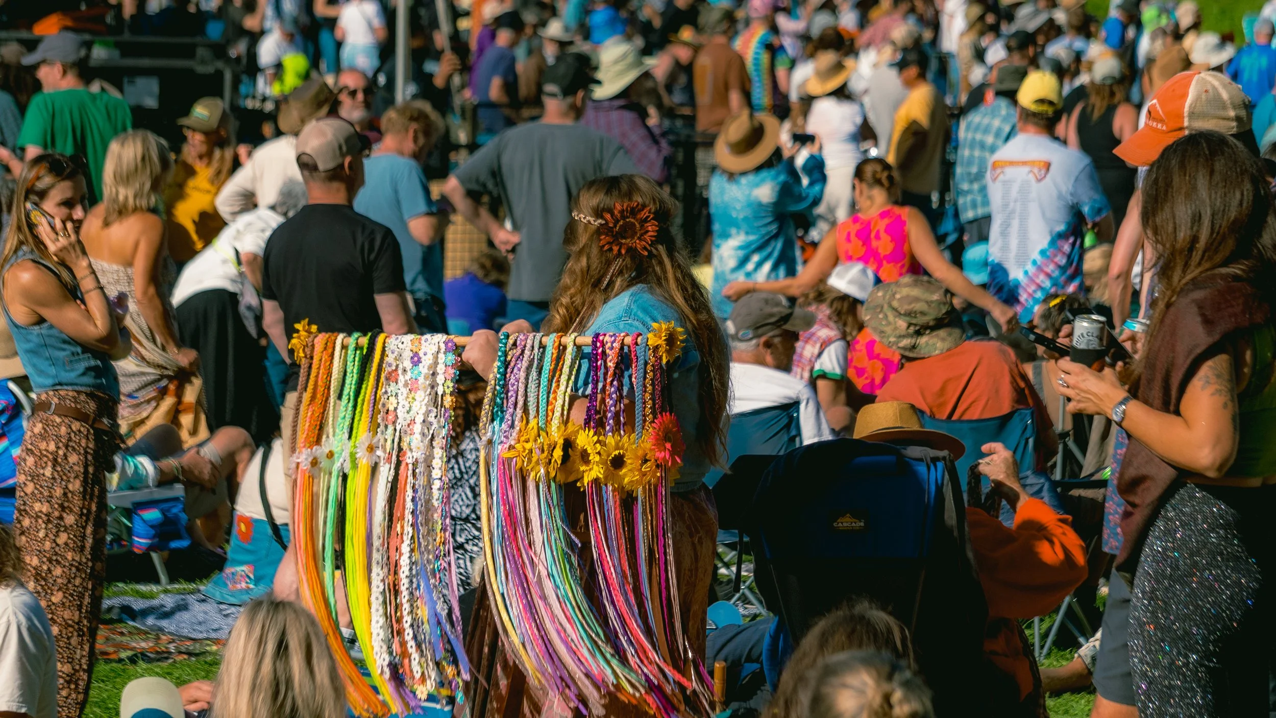 Crowd of people at an outdoor festival or fair, with a woman selling colorful handmade necklaces decorated with sunflowers in the foreground.