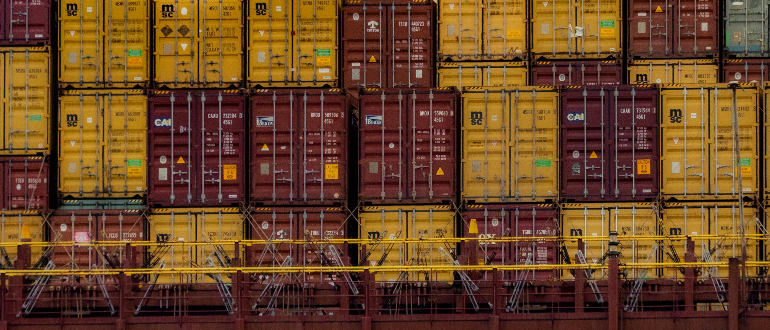 Stacked yellow and red shipping containers on a cargo ship, with metal railings and ladders.