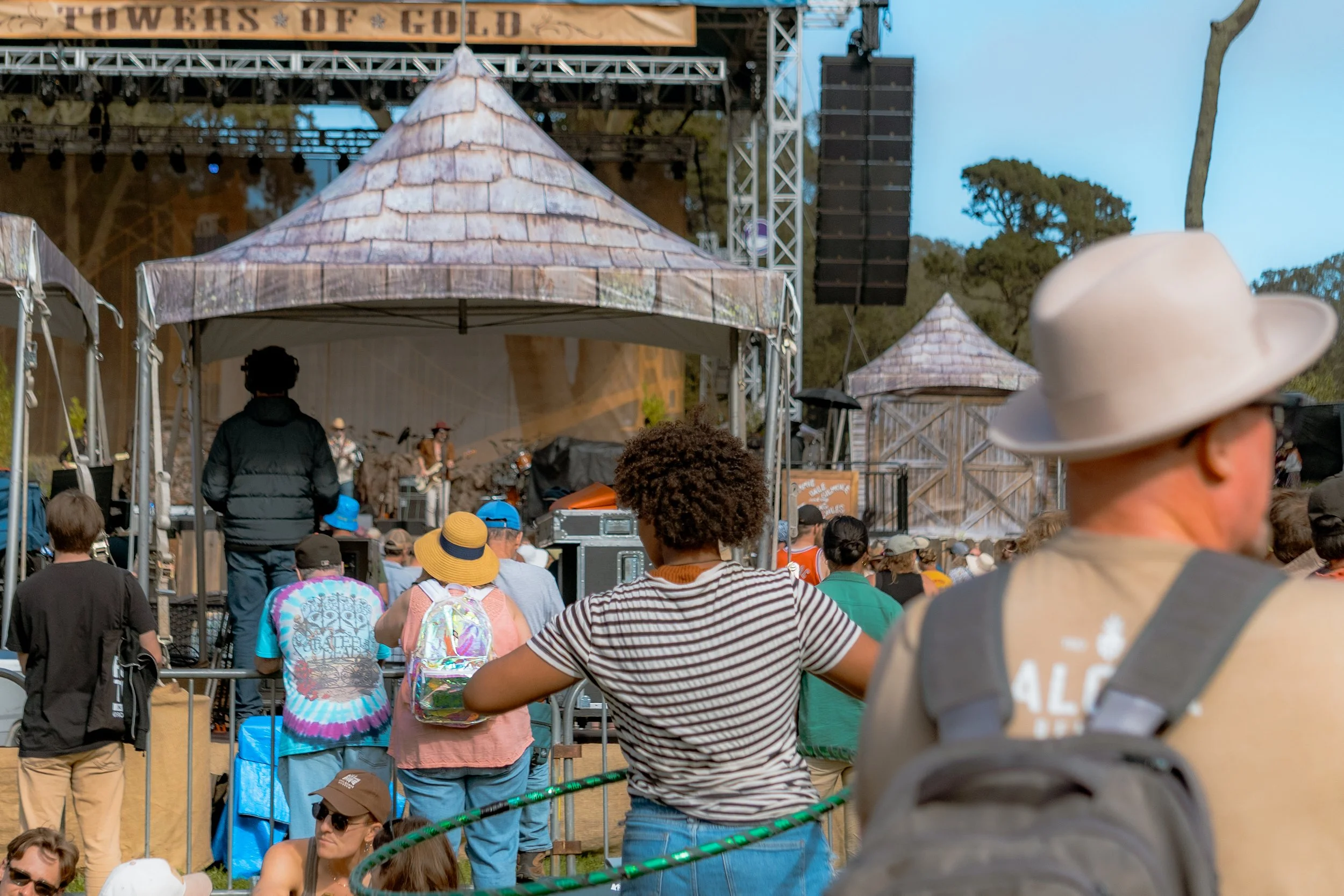 Crowd watching a live outdoor concert with a stage and tents, surrounded by trees.