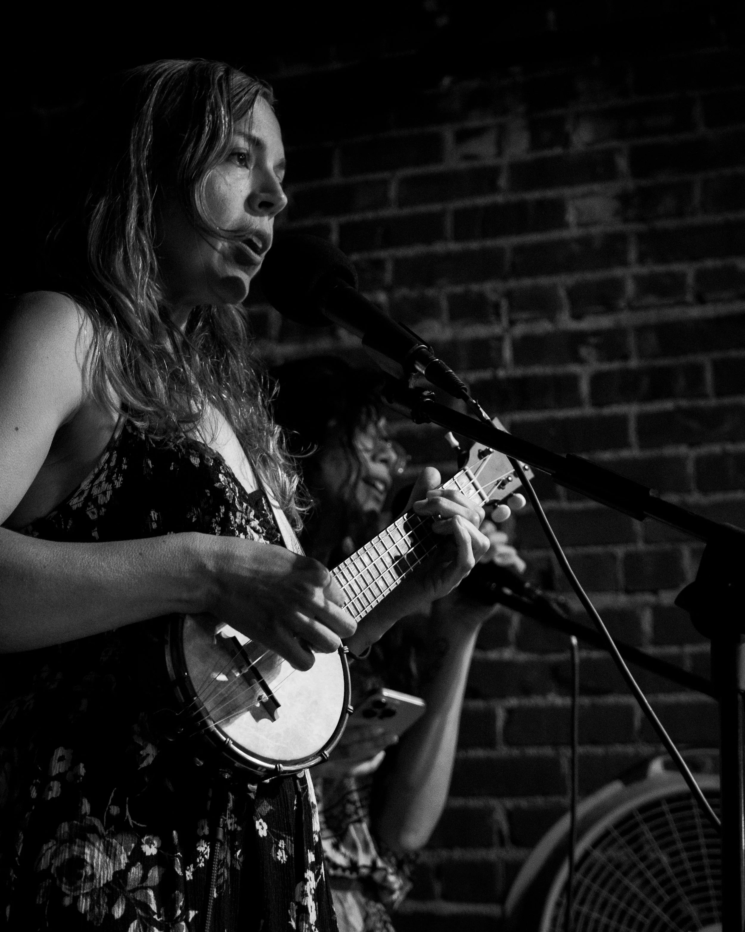 A woman singing into a microphone and playing a ukulele on stage, with a brick wall in the background.