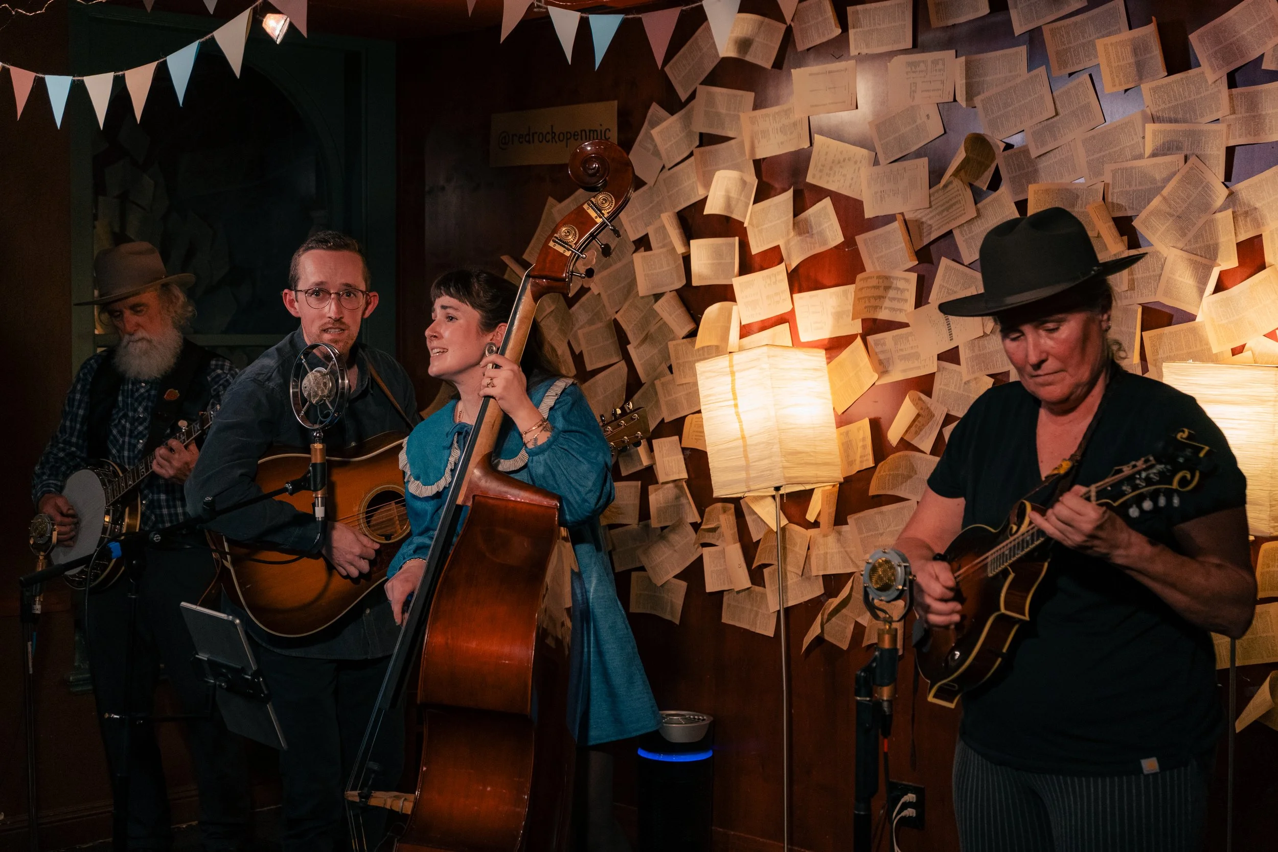 A band playing folk or bluegrass music in a cozy venue with book pages on the wall and warm lighting.