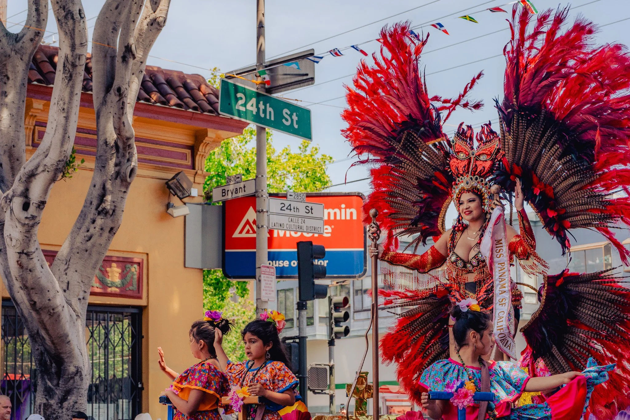 Mexican women and children in colorful traditional costumes participating in a parade on 24th Street in Los Angeles, with a woman dressed in an elaborate red feathered costume, street signs, and a building in the background.