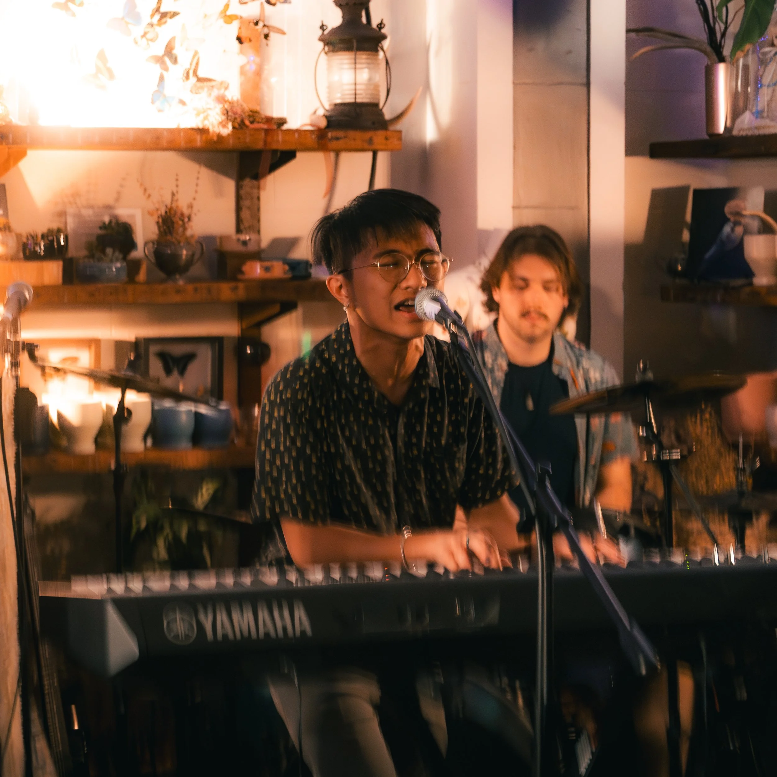 A young man with glasses singing into a microphone while playing a Yamaha keyboard, with a drummer in the background, in a cozy indoor setting with warm lighting and decorative shelves.
