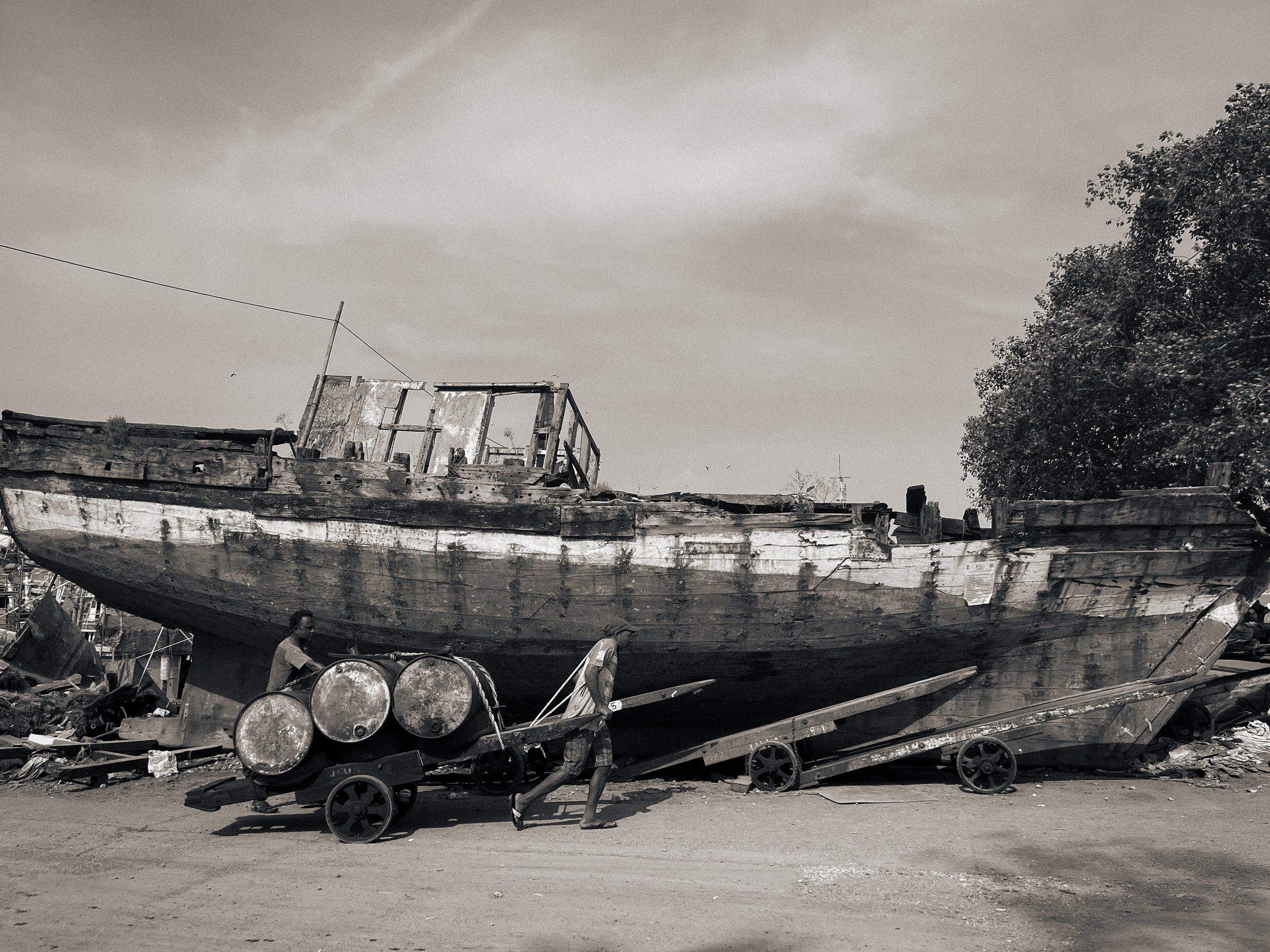Two people pushing a cart with four large barrels past a giant, weathered, abandoned boat on land, with a large tree and cloudy sky in the background.