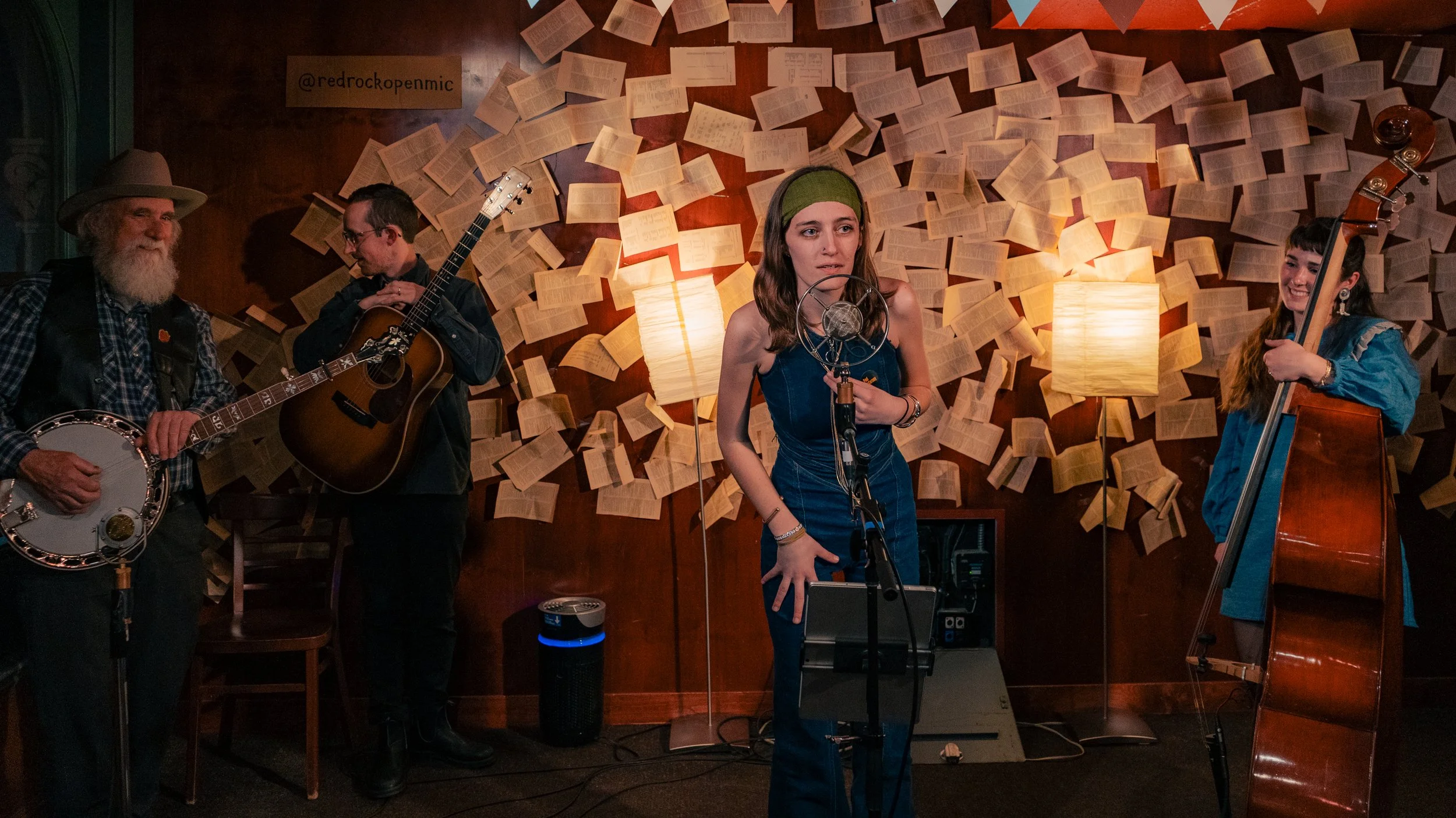 Four musicians performing in a cozy indoor setting with a background of open books and warm lighting. A man with a grey beard wearing a hat plays a banjo, another man next to him plays an acoustic guitar, a woman in a blue dress with a green headband
