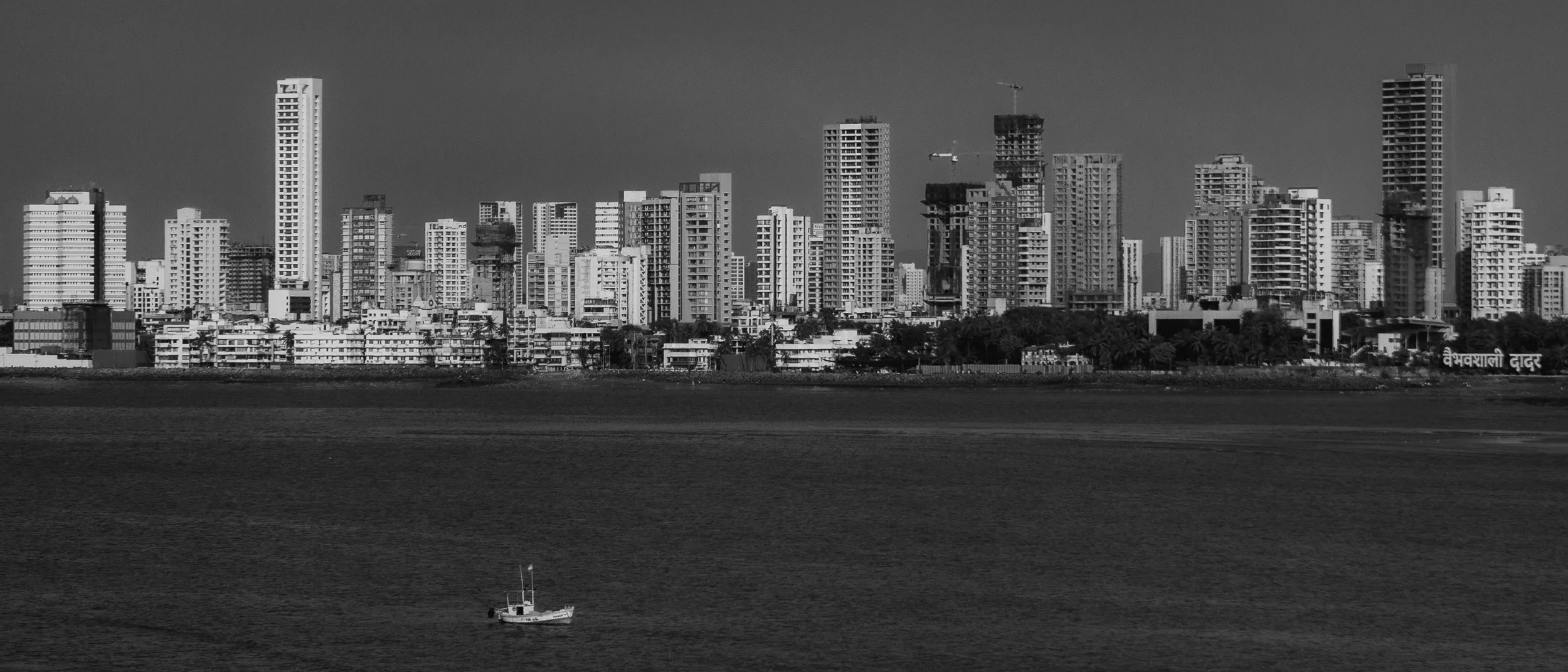 Black and white photo of a city skyline with tall buildings across a body of water, with a small boat in the foreground.