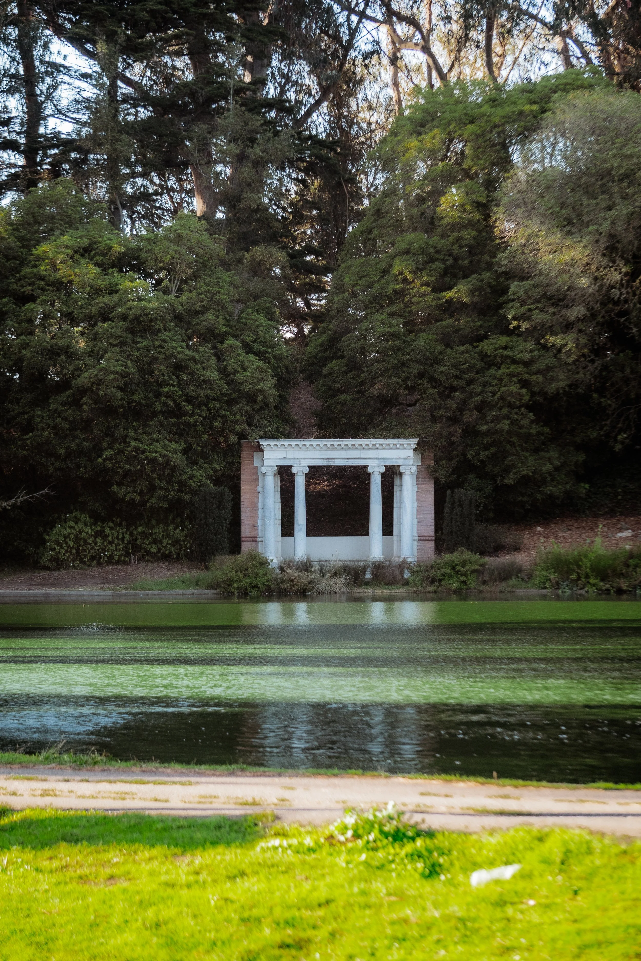 A white, classical-style pavilion with columns, situated behind a pond with green algae, surrounded by trees and lush greenery.