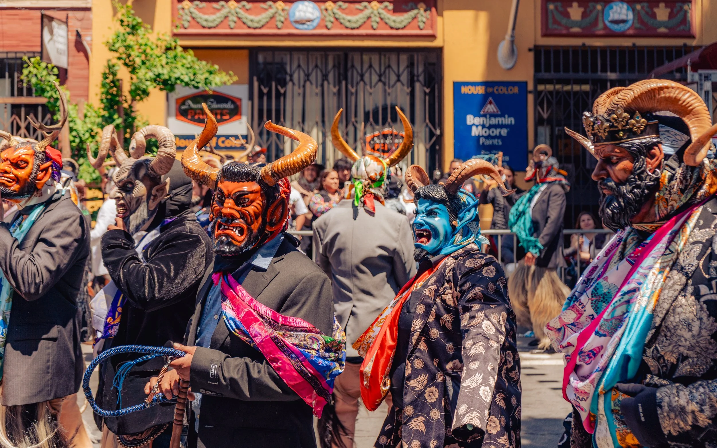 Men dressed in colorful costumes and masks with horns, participating in a street parade or festival, with onlookers in the background.