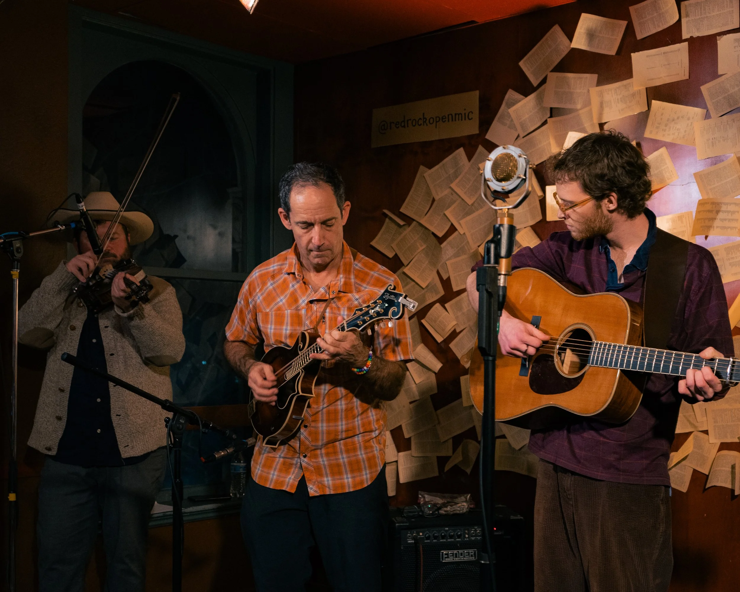 Three musicians performing live in a recording studio, with one man playing a mandolin, another playing an acoustic guitar, and a third holding a camera, surrounded by pages of sheet music on the wall.