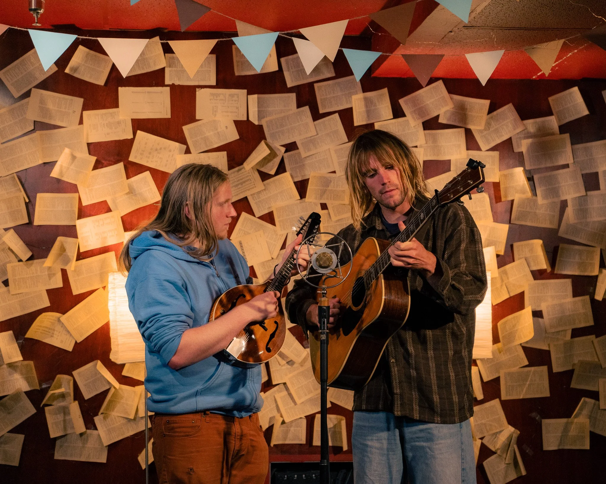 Two musicians playing guitars, one on a mandolin, performing in a cozy room decorated with papers and bunting, with warm lighting.