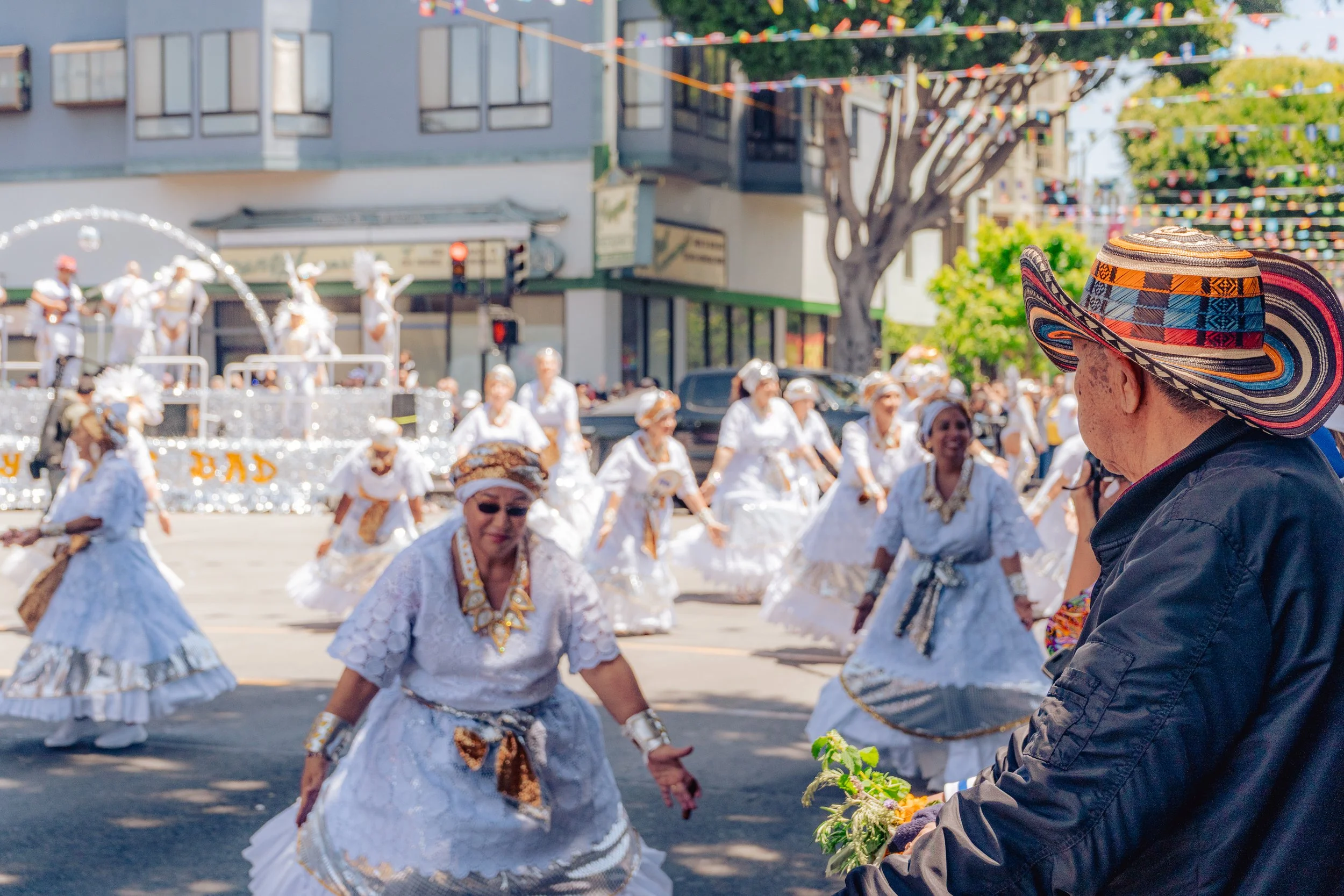 A street parade with a group of women dressed in traditional white folkloric dresses dancing, while an older man wearing a colorful wide-brimmed hat and black jacket watches and holds flowers. Colorful banners strung across the street and a float wit